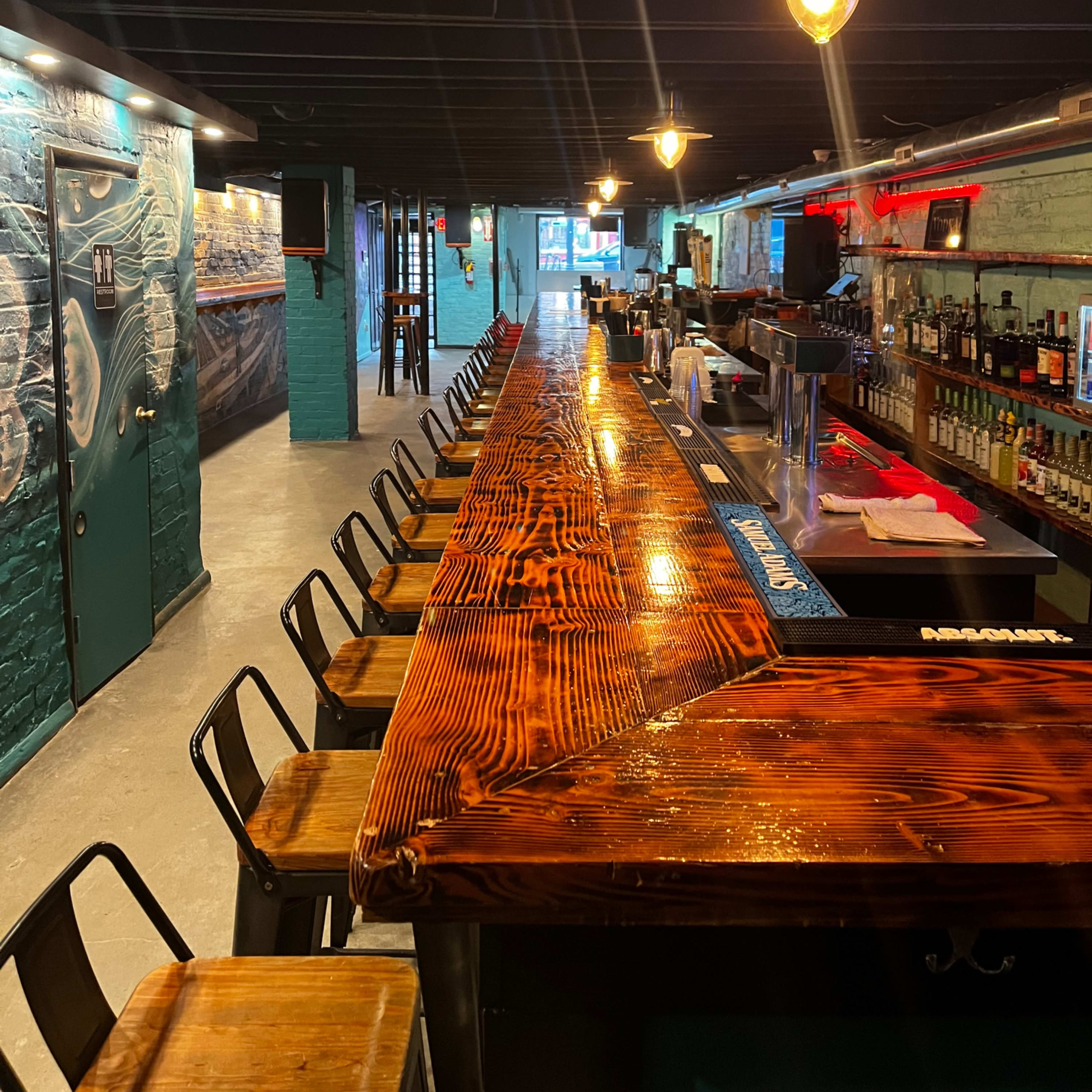 A dimly lit bar with a long wooden countertop and several stools lined up along one side, leading to a well-stocked bar area in the background.