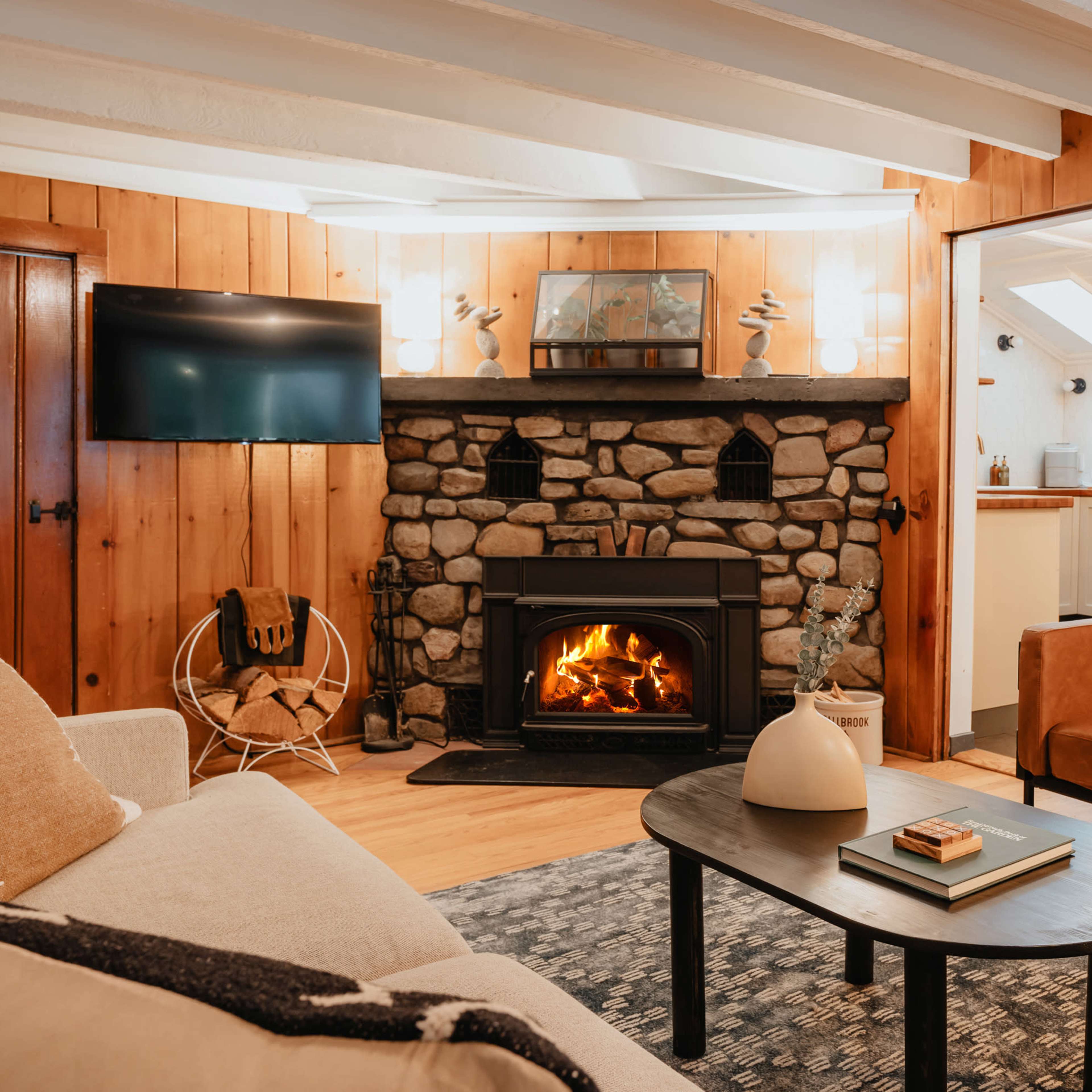 A cozy living room with wooden walls, a stone fireplace, a television mounted on the wall, and two armchairs arranged around a coffee table.