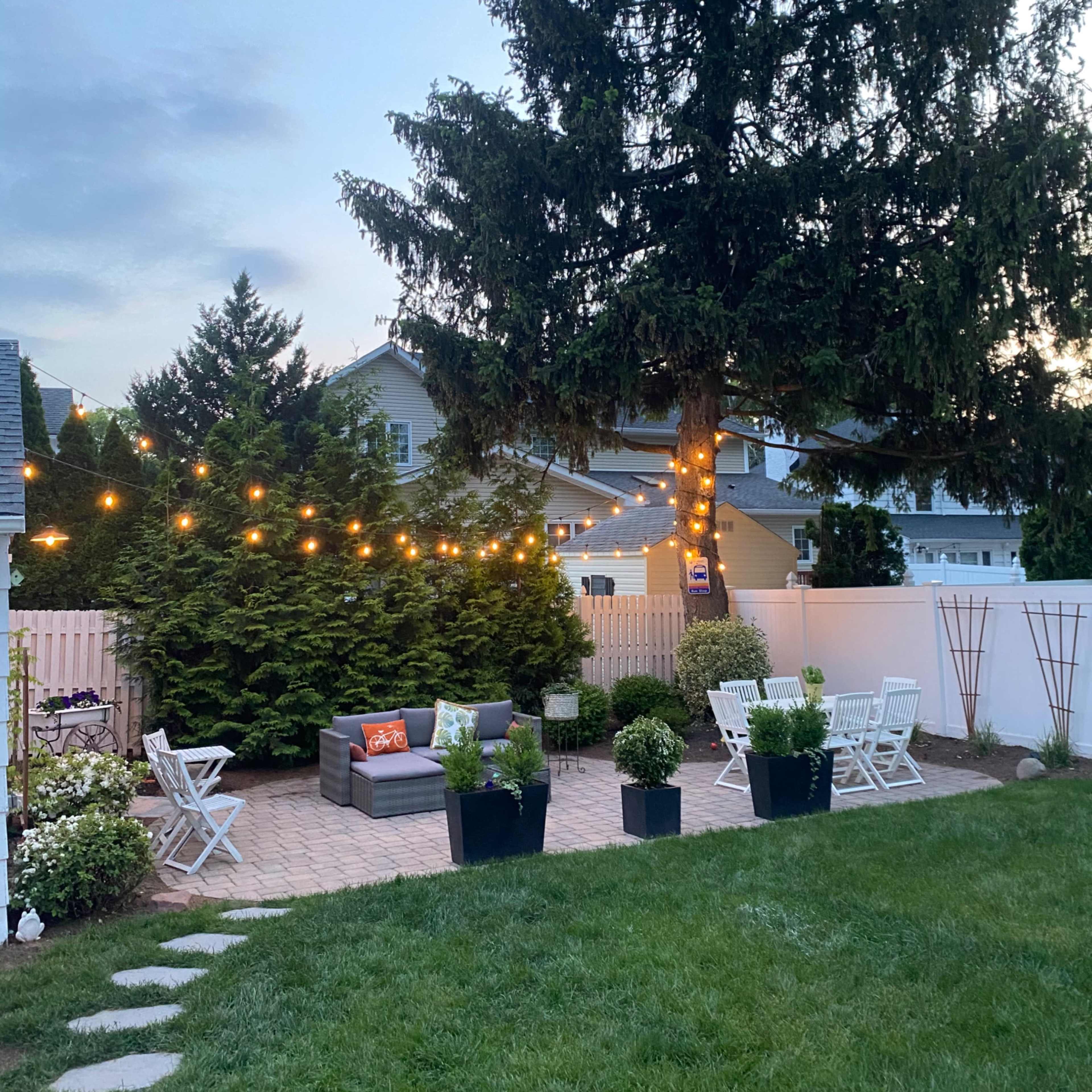 A backyard patio area with string lights overhead, a seating arrangement on a stone patio, and potted plants along the perimeter.