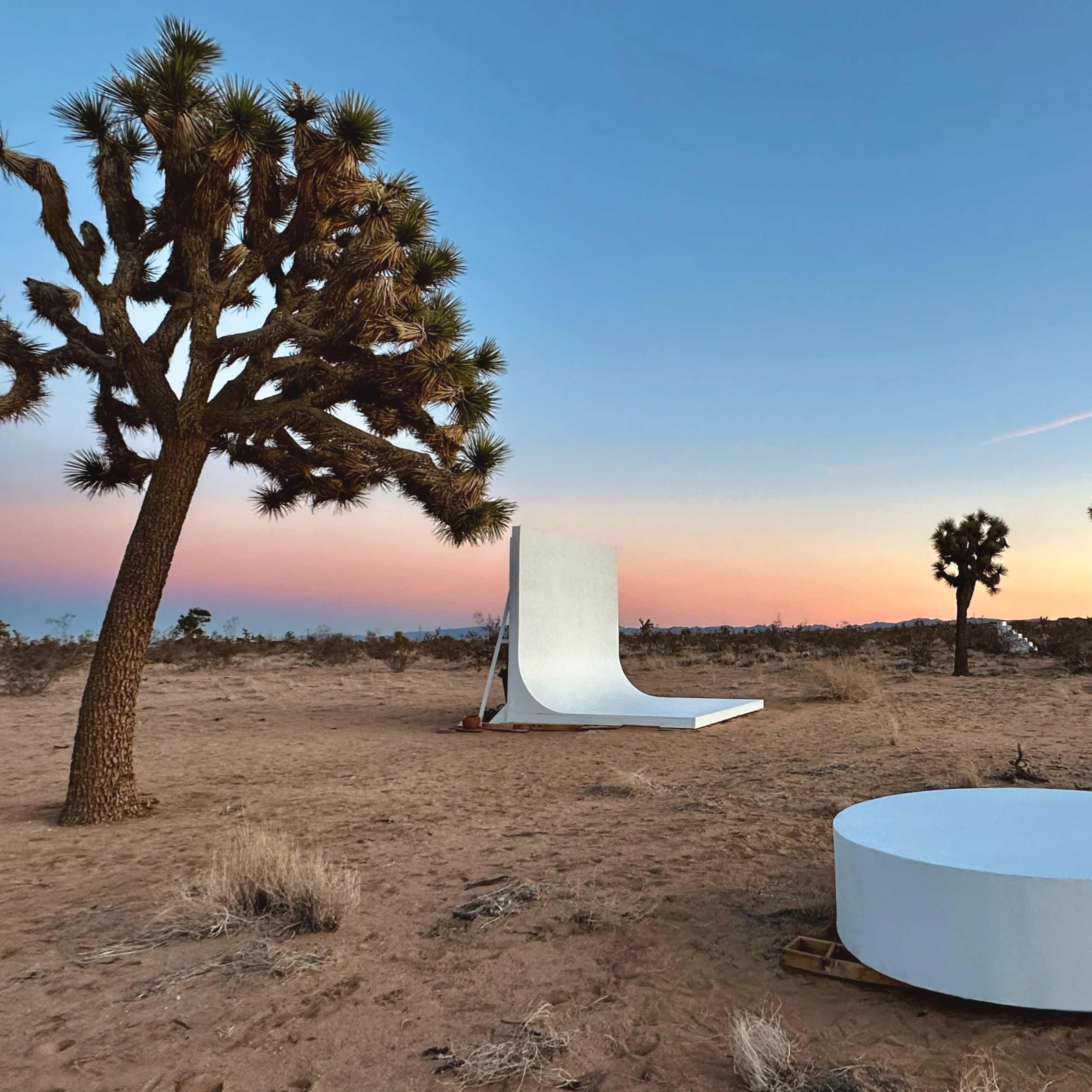 A desert landscape at sunset, featuring a large white backdrop and a circular white platform surrounded by Joshua trees.