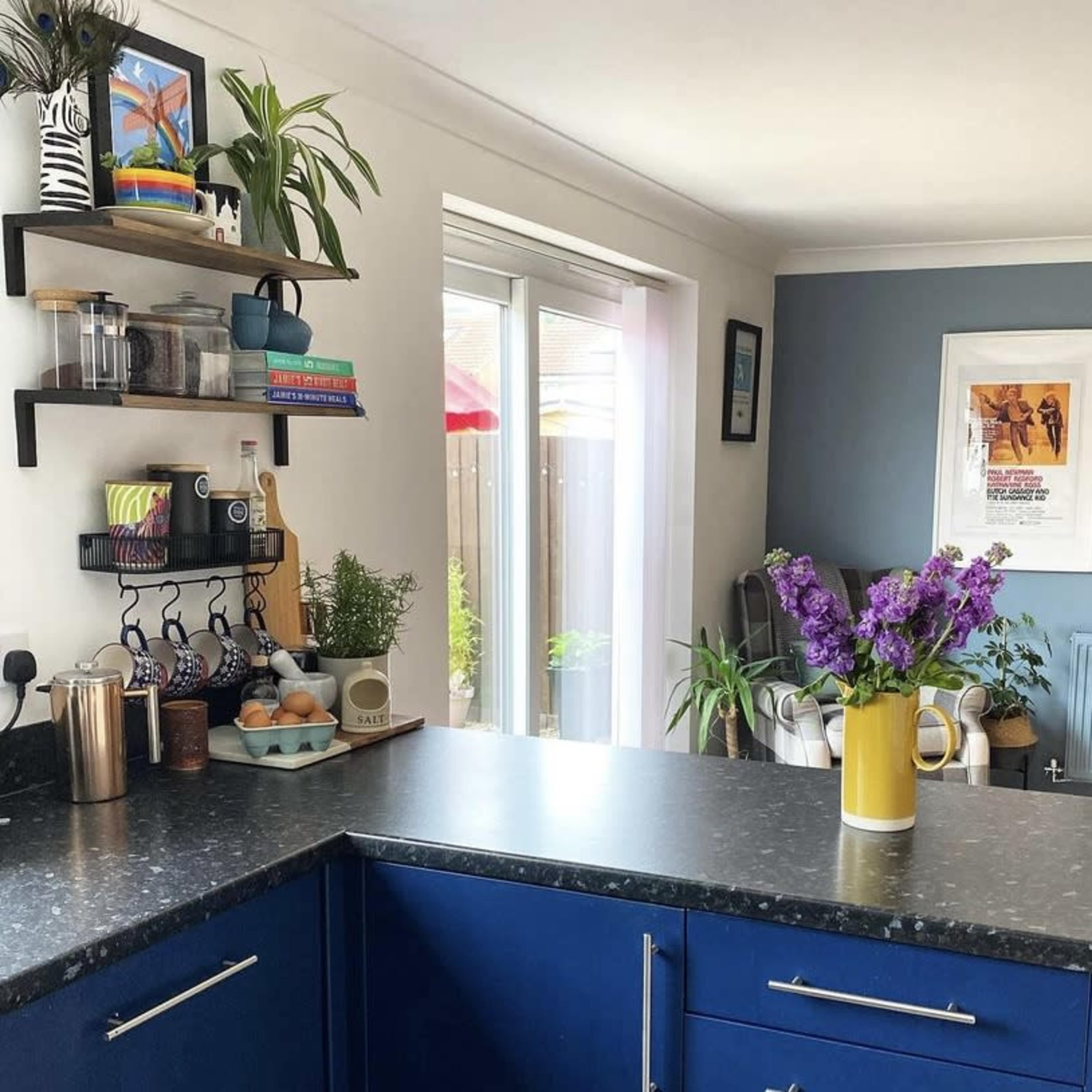 A modern kitchen features a blue countertop, shelves with various kitchen items, and a vase of purple flowers.