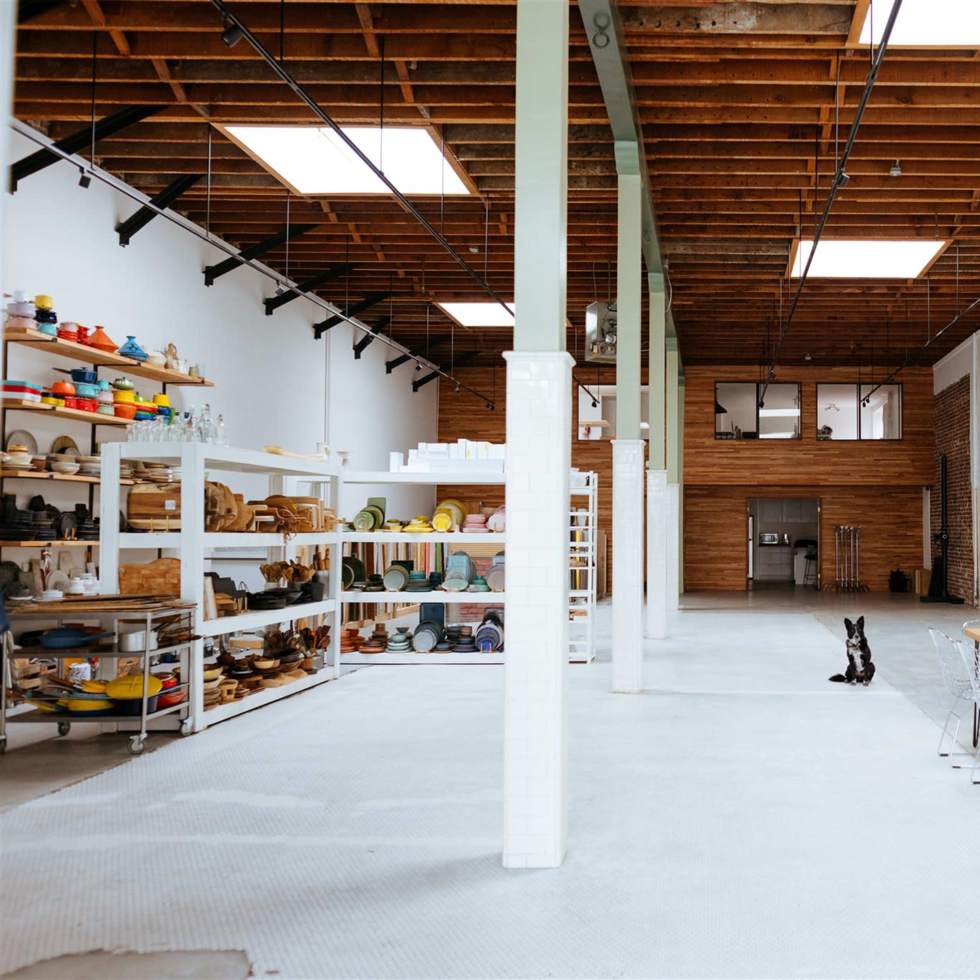 A spacious, well-lit interior features a long wooden table, a collection of colorful pottery on shelves, and a dog sitting on the concrete floor.