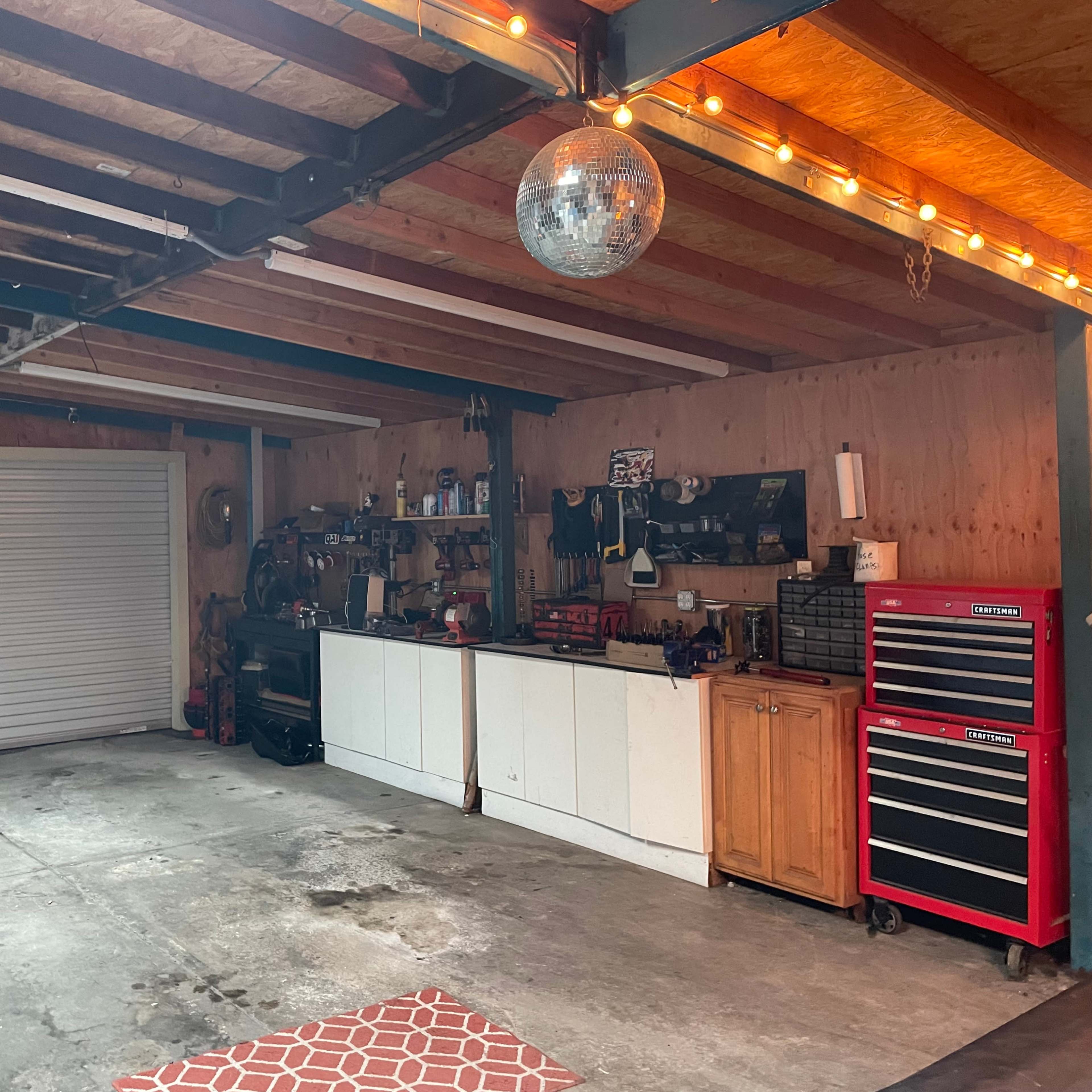 A garage with a roll-up door, a workbench against one wall, and two tool chests, all illuminated by string lights overhead.