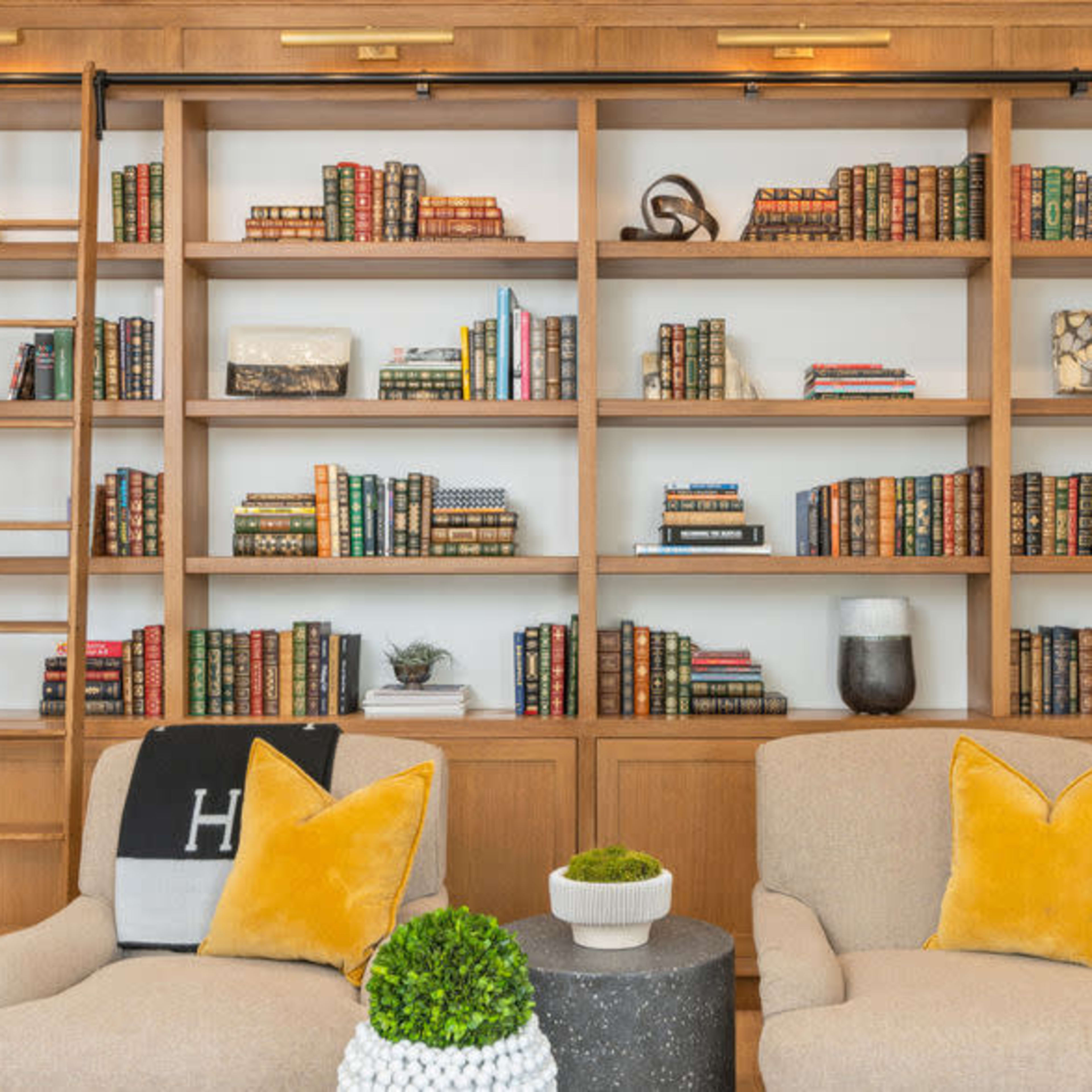 A wooden shelving unit filled with neatly arranged books and decorative items, flanked by two upholstered chairs and a ladder.