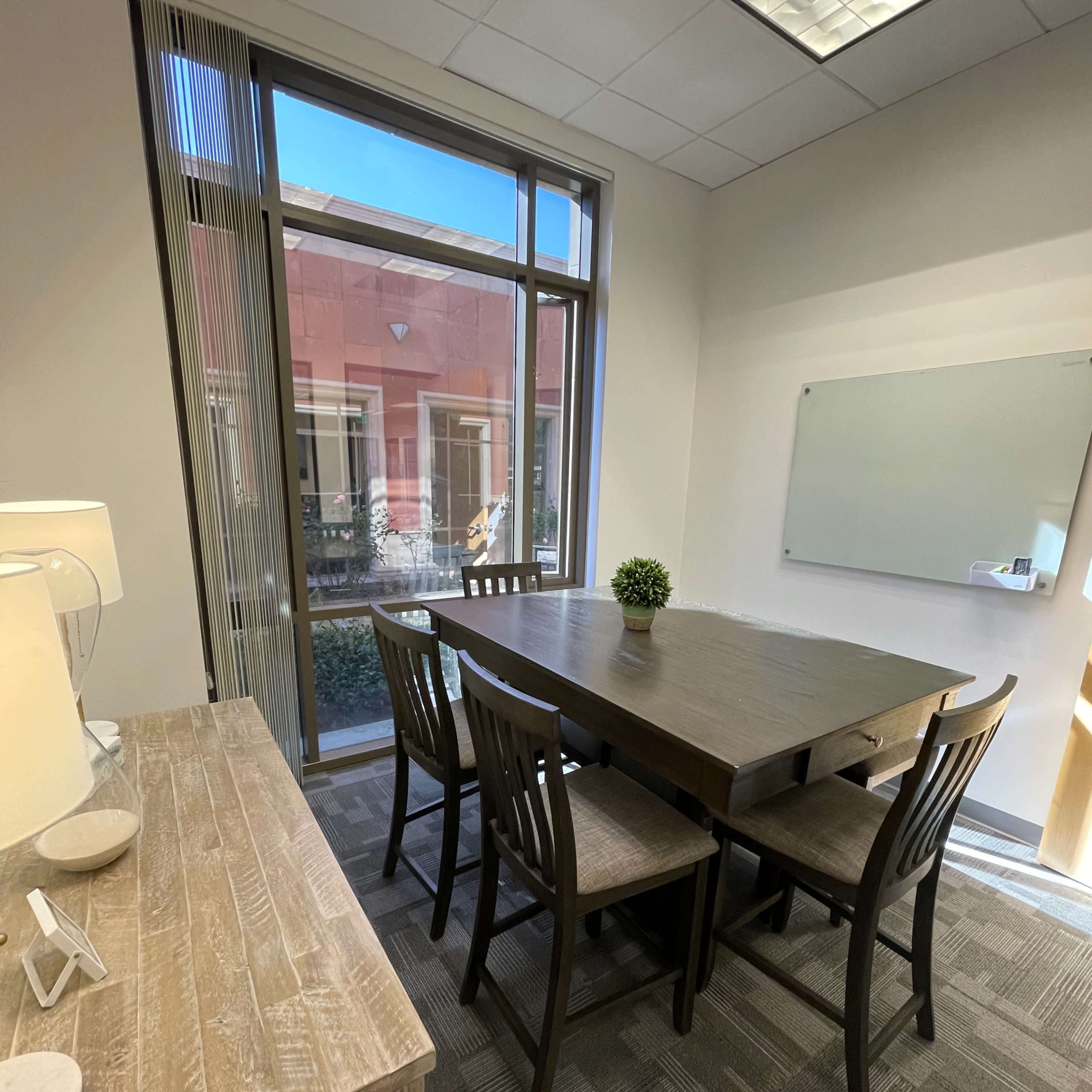 A modern conference room features a wooden table surrounded by four chairs, with a plant centerpiece and a whiteboard on the wall.