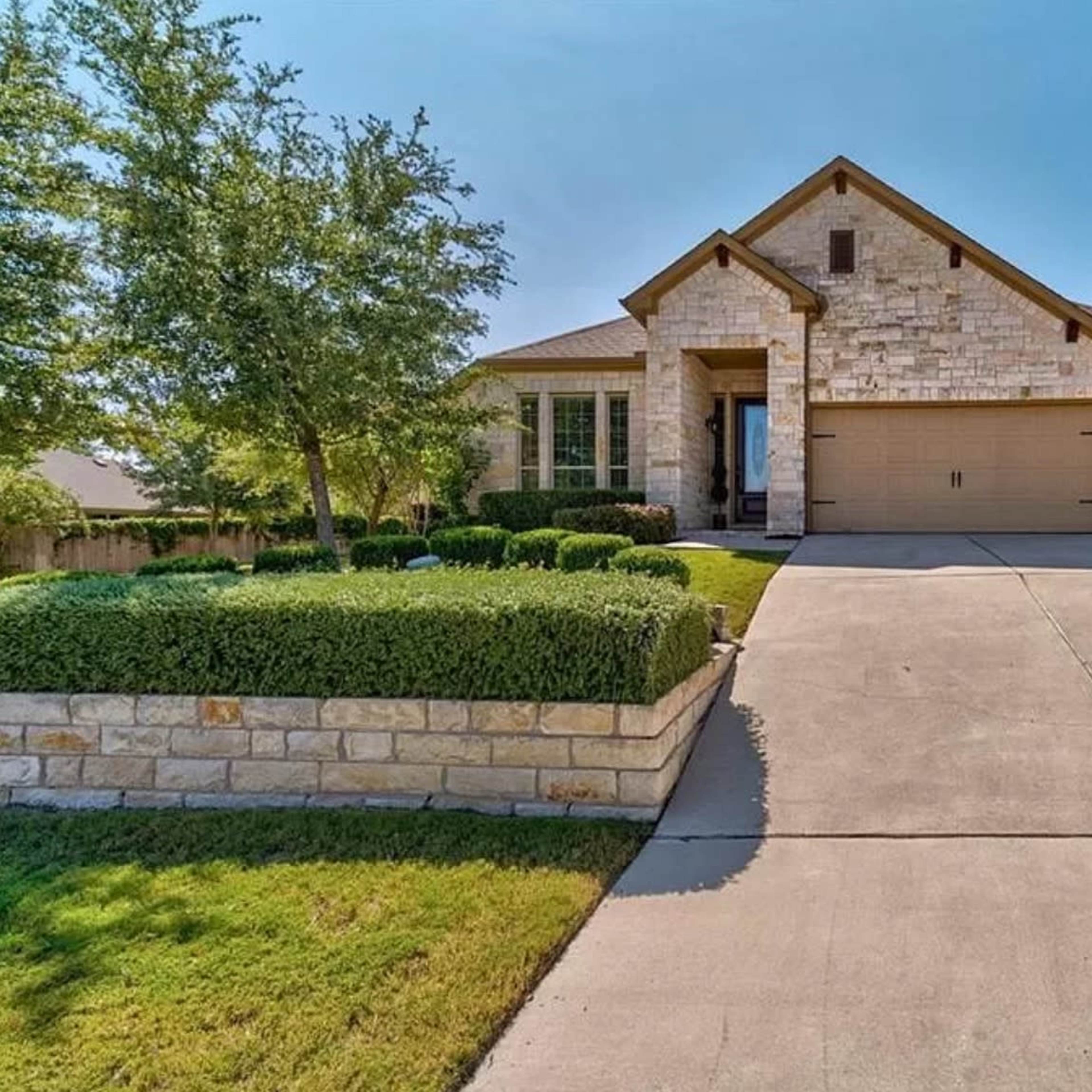 A single-story stone house with a two-car garage sits on a large concrete driveway surrounded by neatly trimmed shrubs and trees.