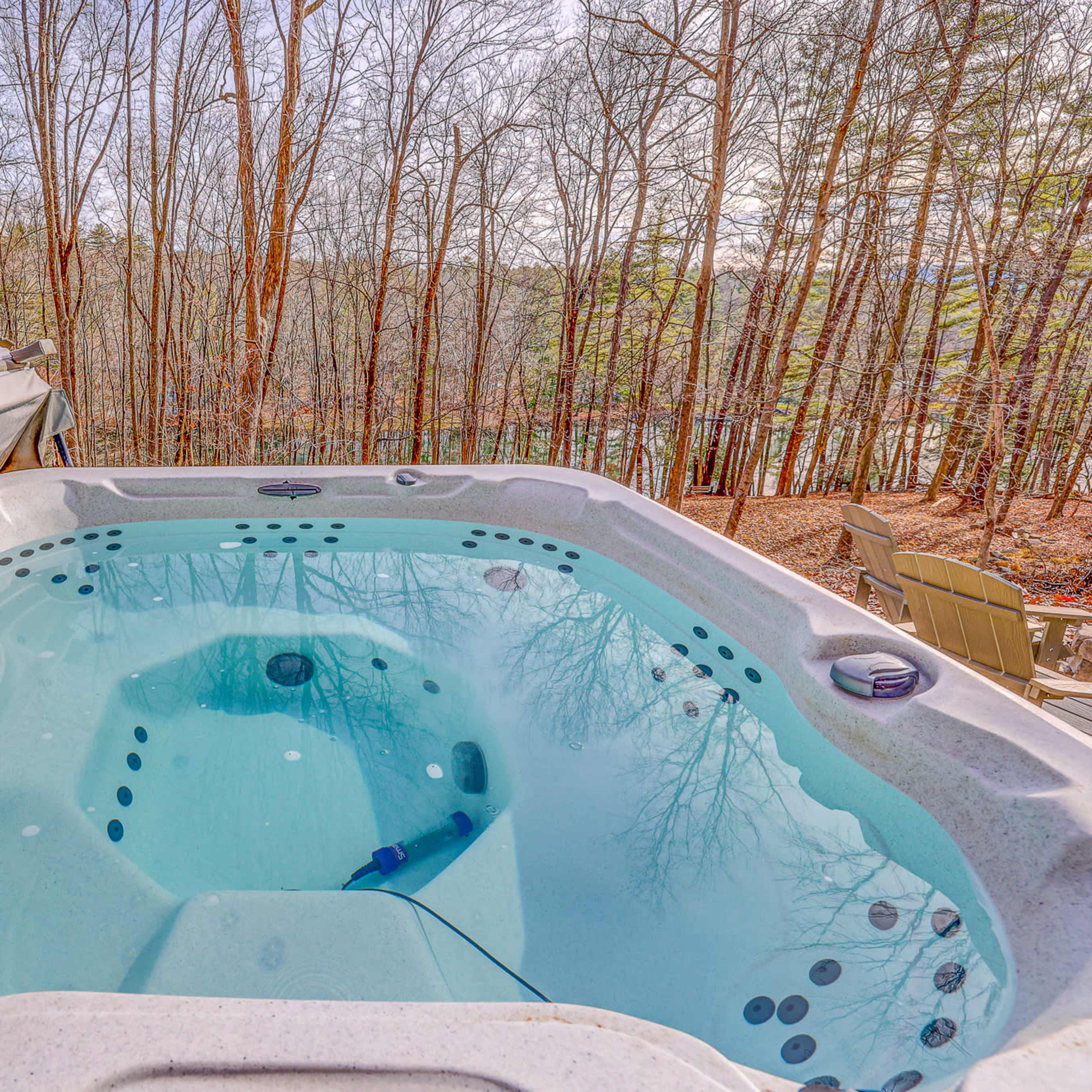 A hot tub is set on a deck overlooking a wooded area with bare trees and a calm body of water in the background.