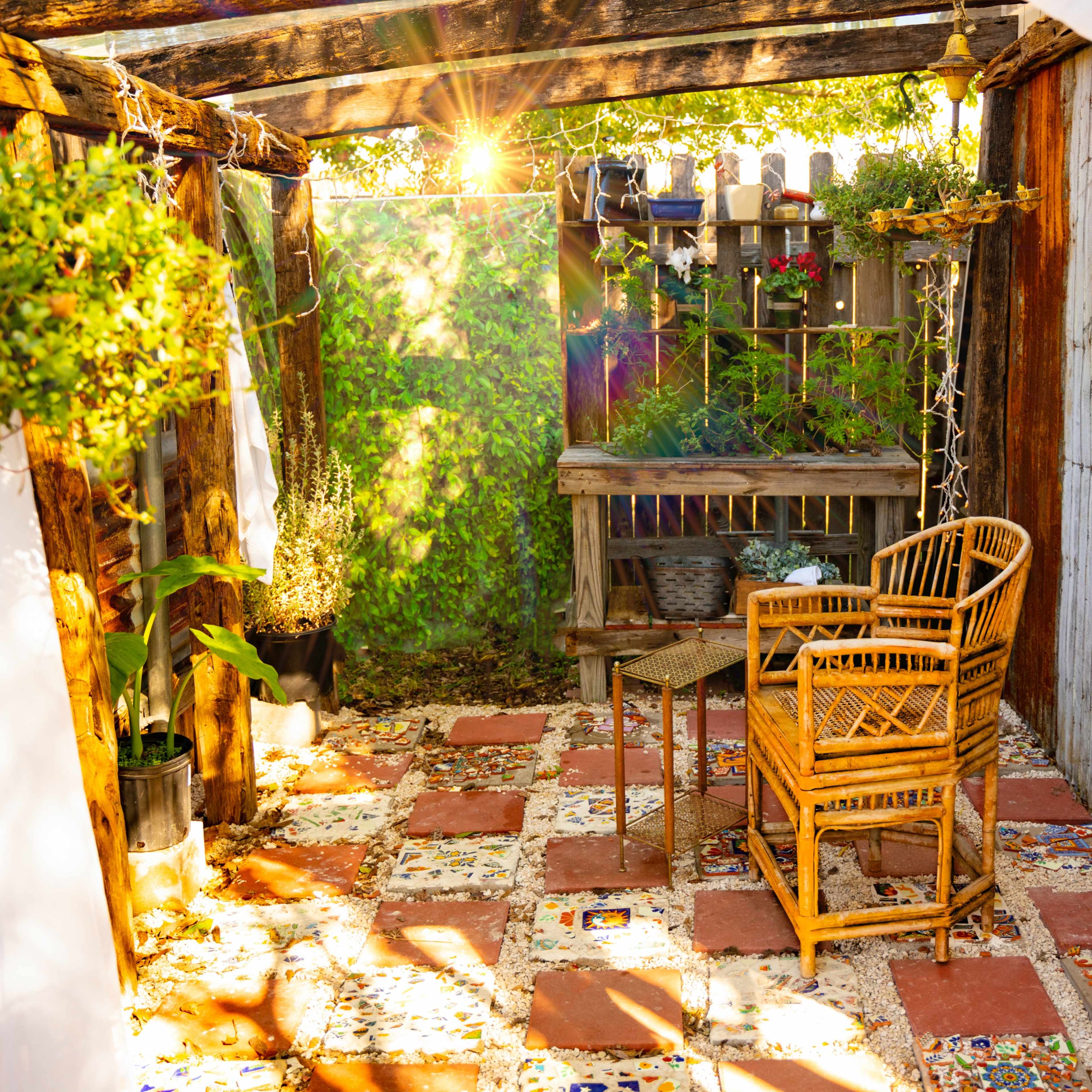 A cozy outdoor seating area features a rattan chair and small table surrounded by greenery and colorful tiled flooring.