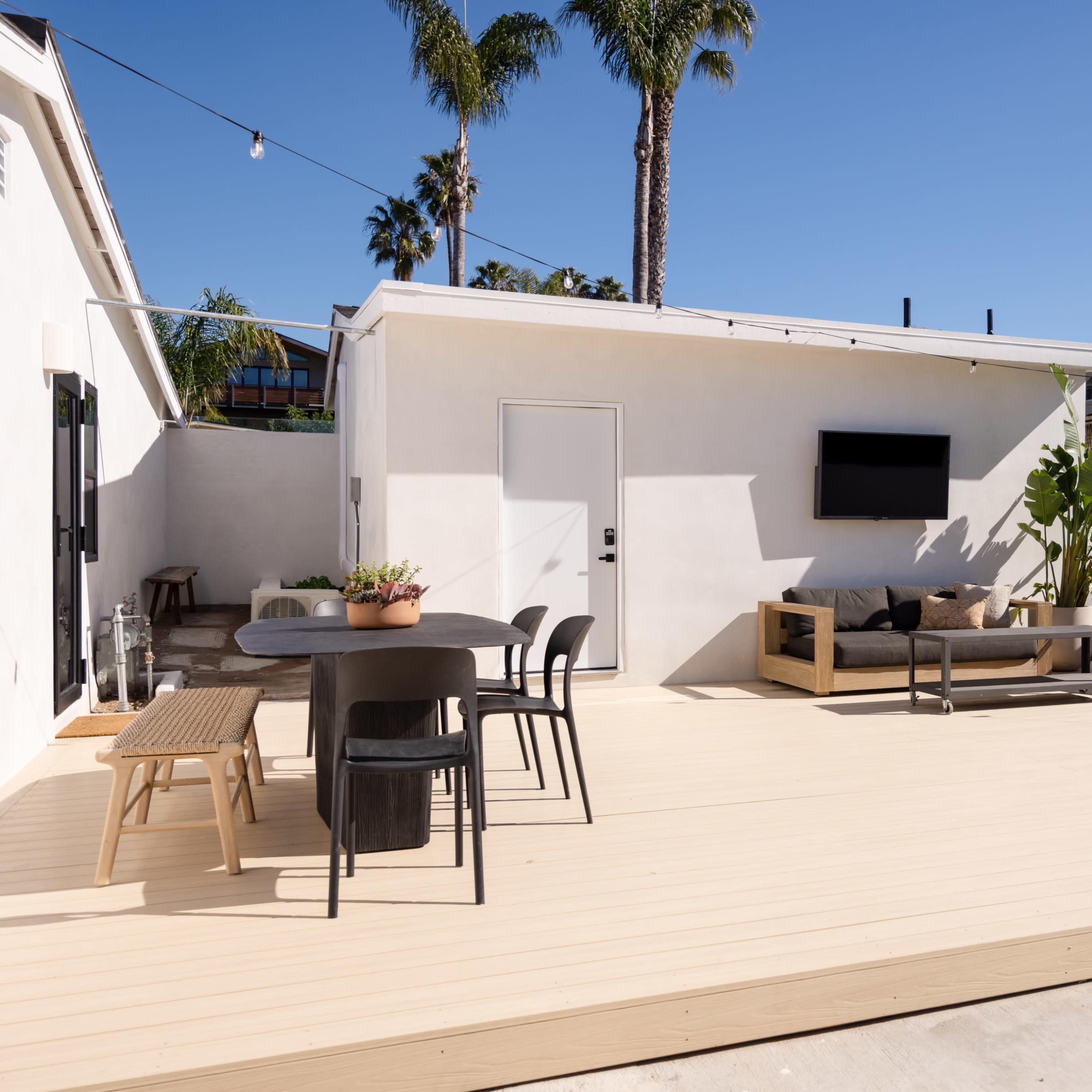 A modern outdoor patio area featuring a wooden deck, a dining table, lounge seating, and palm trees under a clear blue sky.