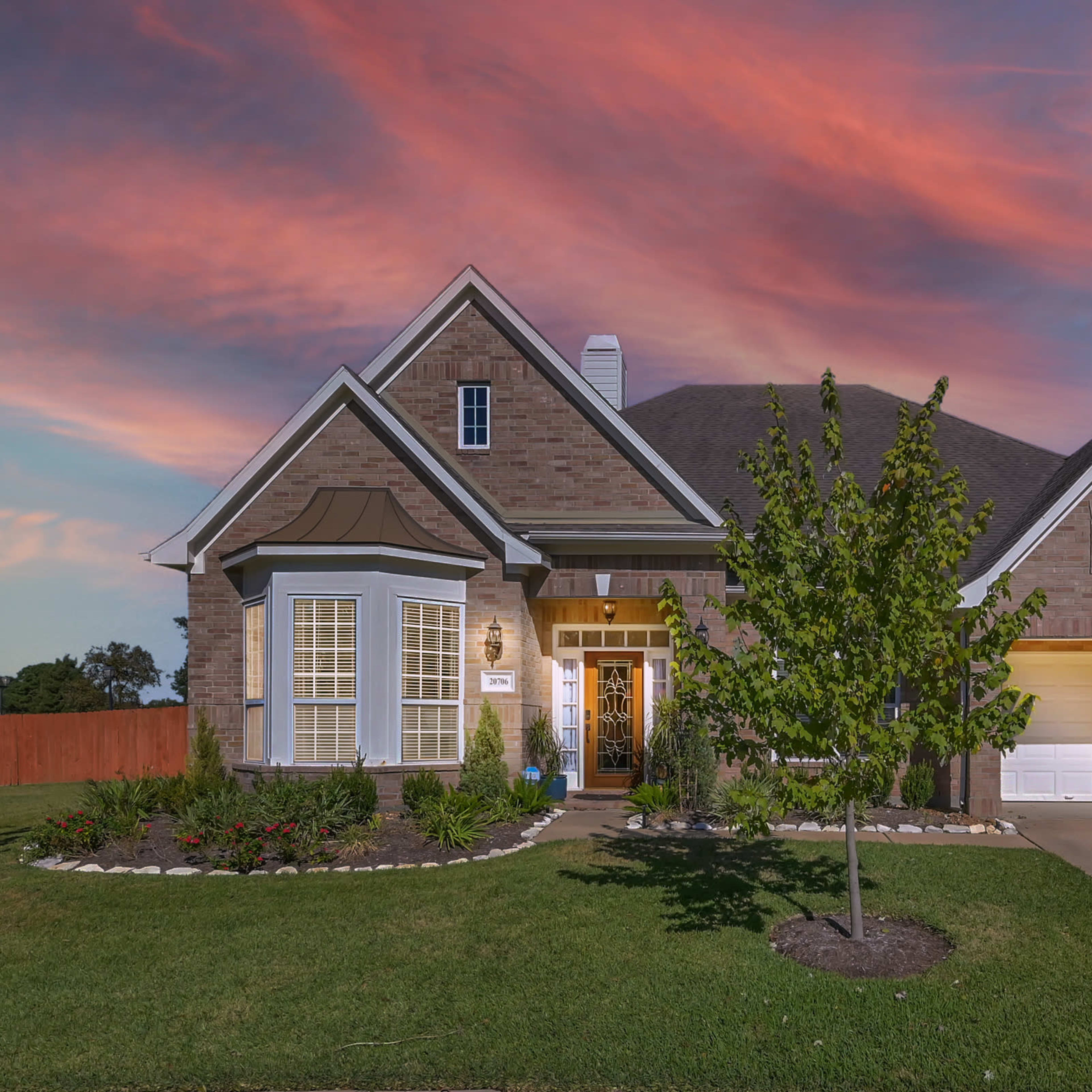 A single-story brick house with a manicured lawn and a driveway leading to two garages is illuminated under a colorful sunset sky.