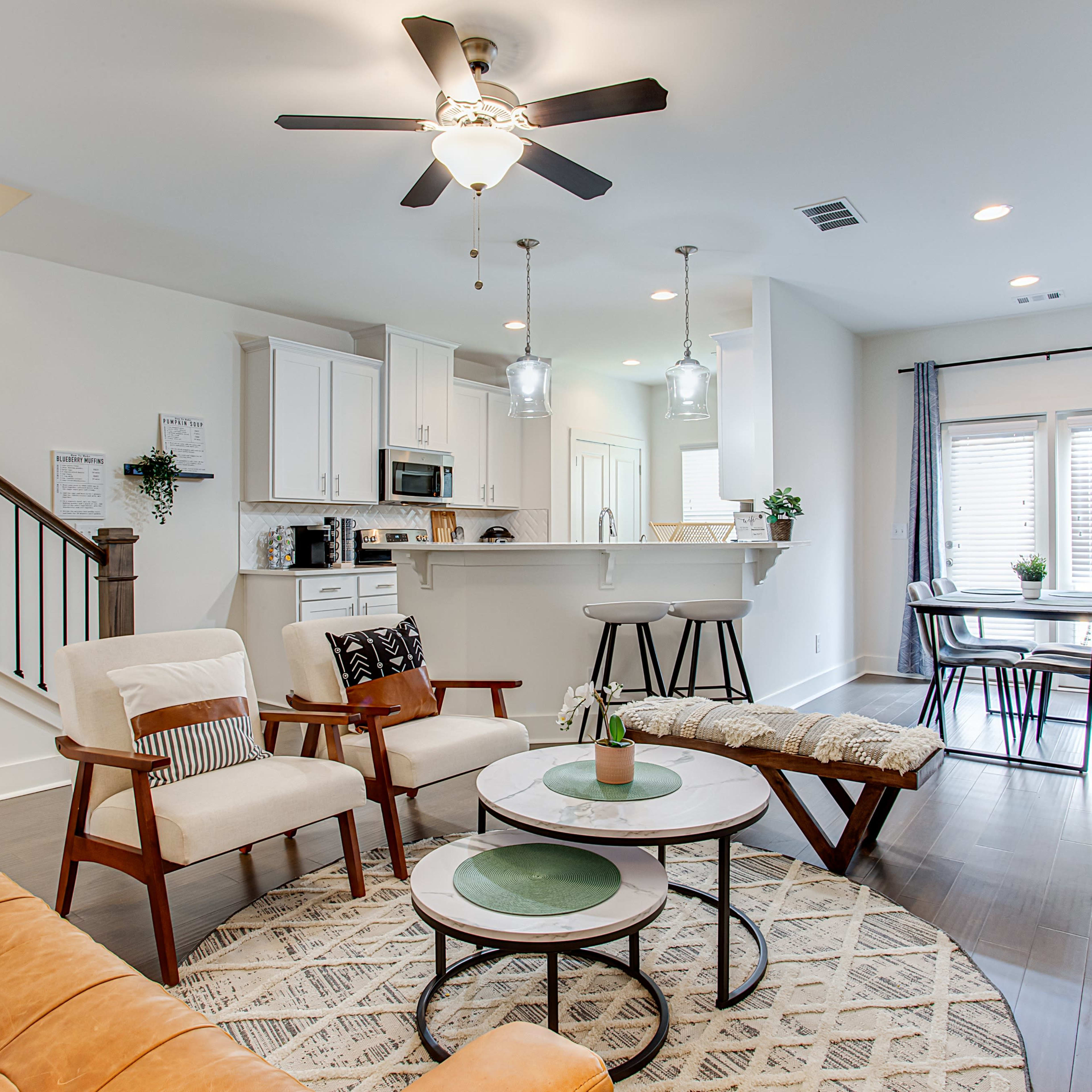 A modern living room features a tan couch, white chairs, a round coffee table, and a dining area in the background with a table and chairs.