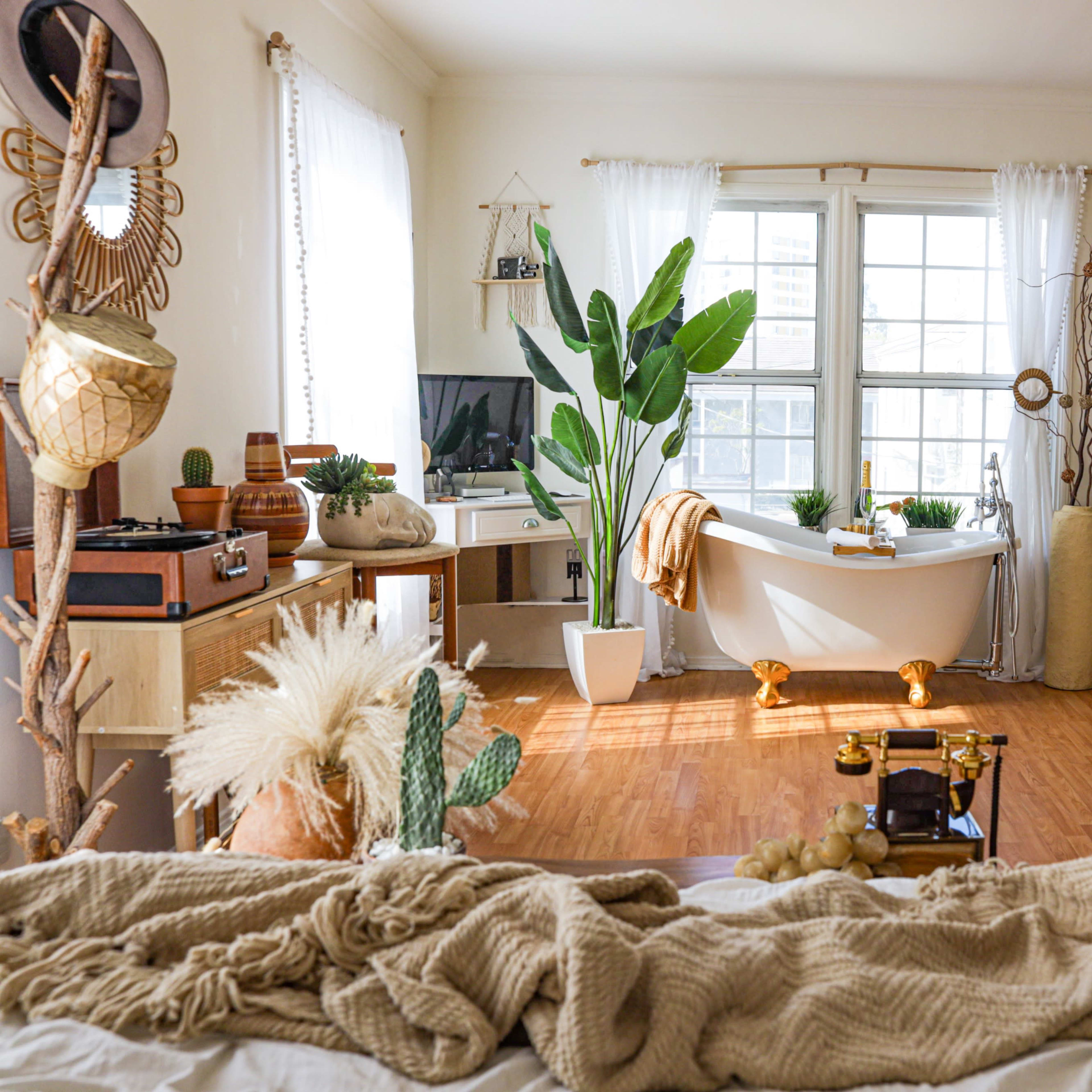 The bright living space features a white bathtub, large windows, several plants, and various decorative items arranged around wooden flooring.