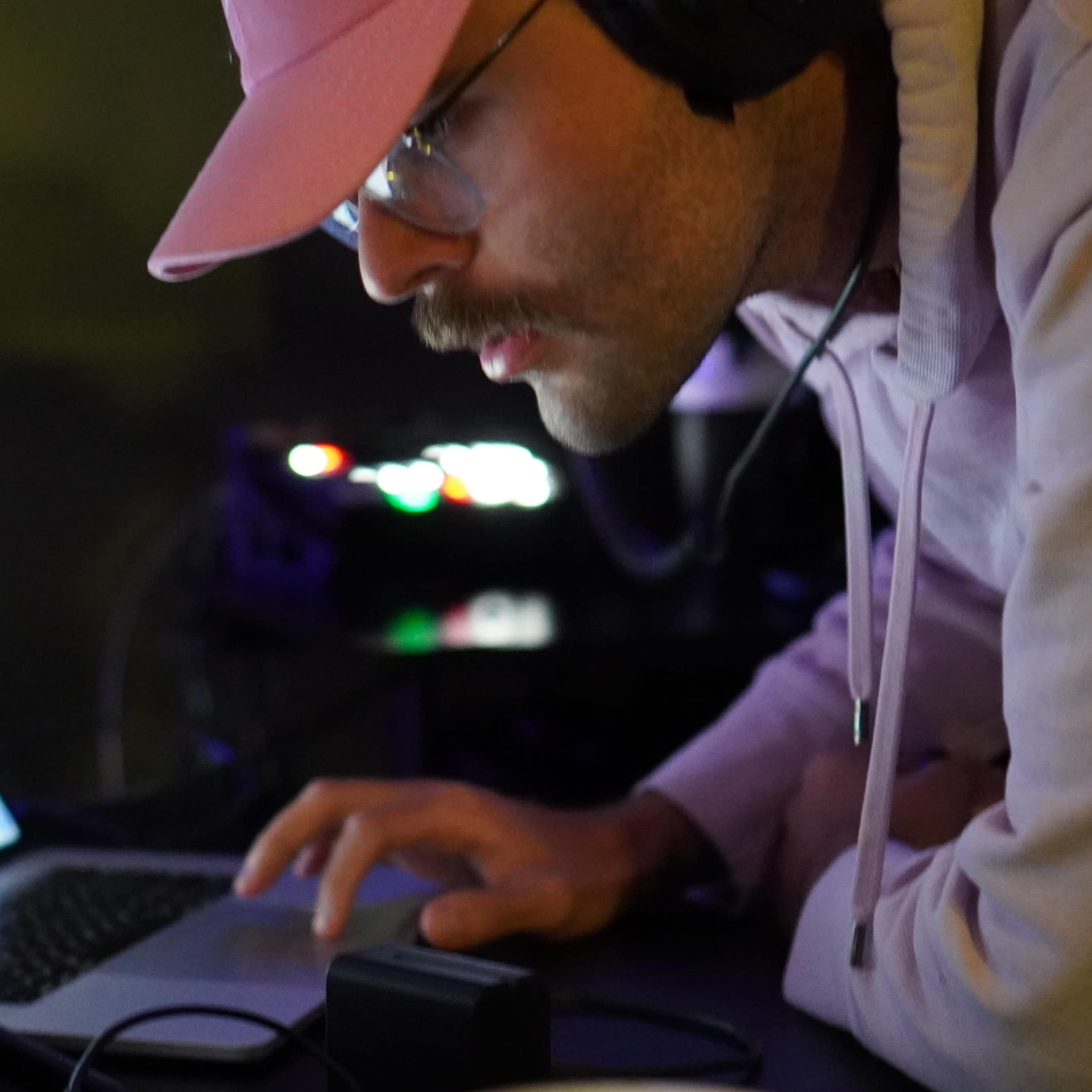 A man wearing a pink hat and glasses leans over a laptop while adjusting settings on a tablet in a dimly lit space.