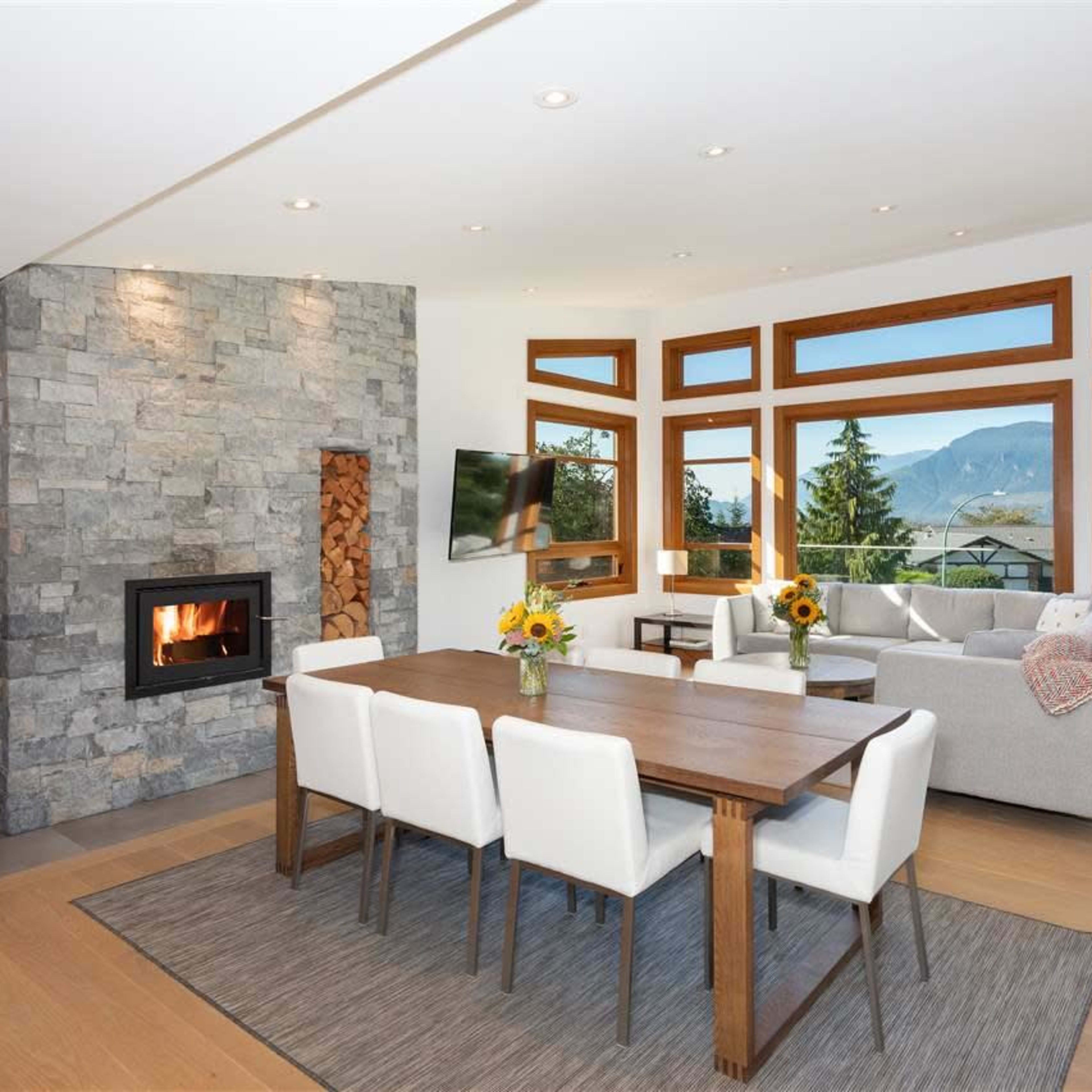 A modern open-concept living and dining area features a stone fireplace, a wooden dining table with white chairs, and large windows that showcase a mountain view.