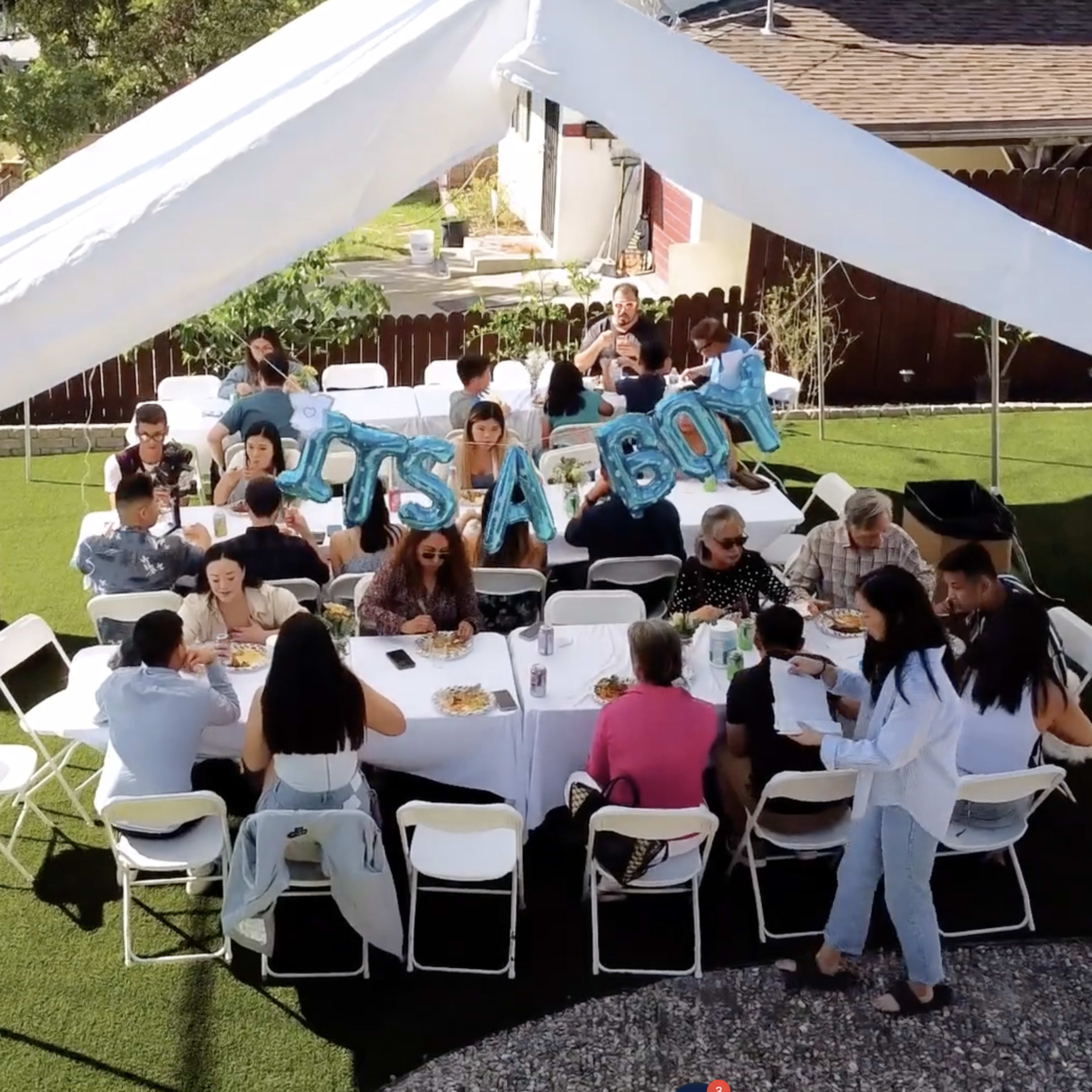 A gathering of people is seated under a white tent with blue "It's a Boy" balloons, celebrating an event in a backyard.