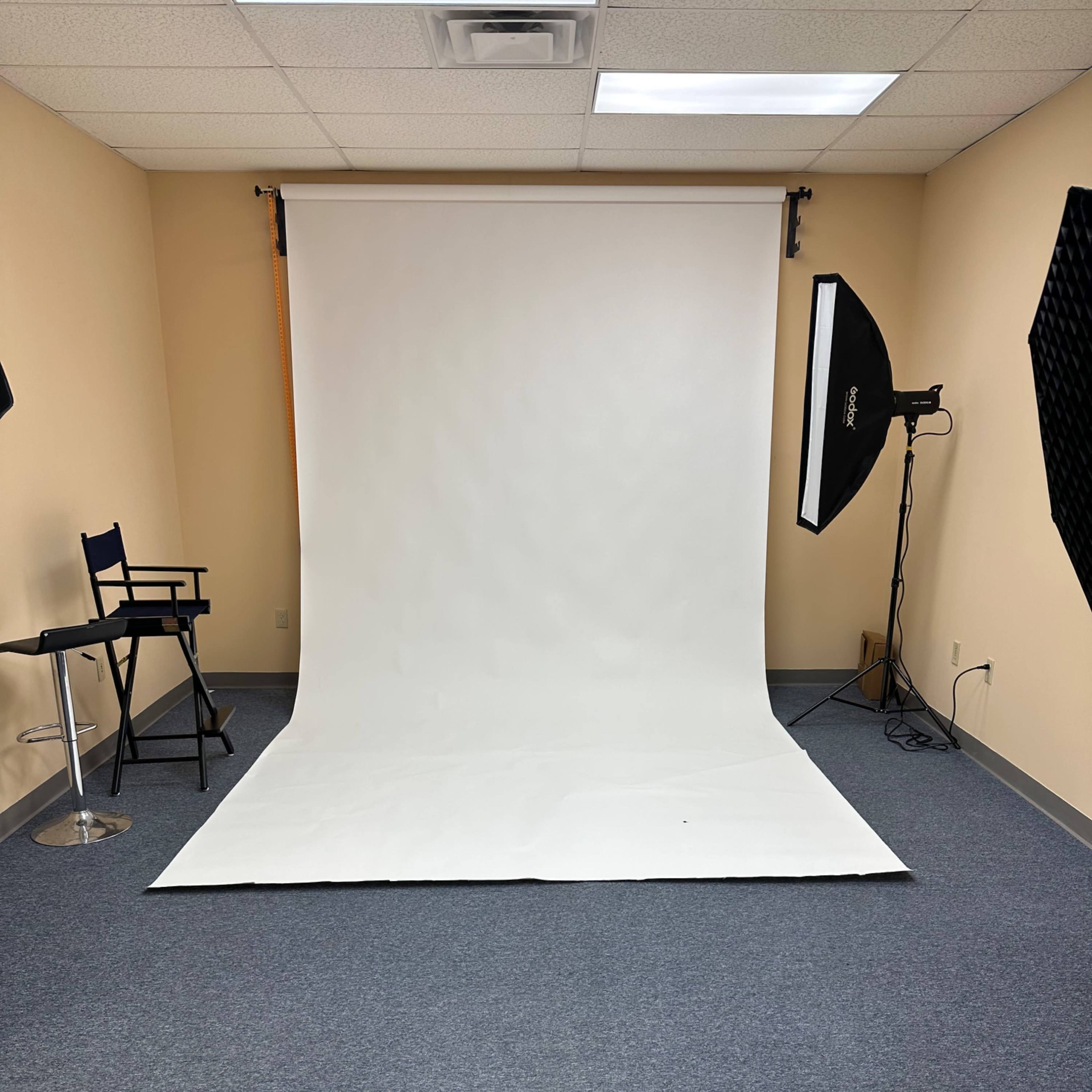 A photography studio with a white backdrop, a chair, and studio lighting equipment set up in a room with gray carpet.