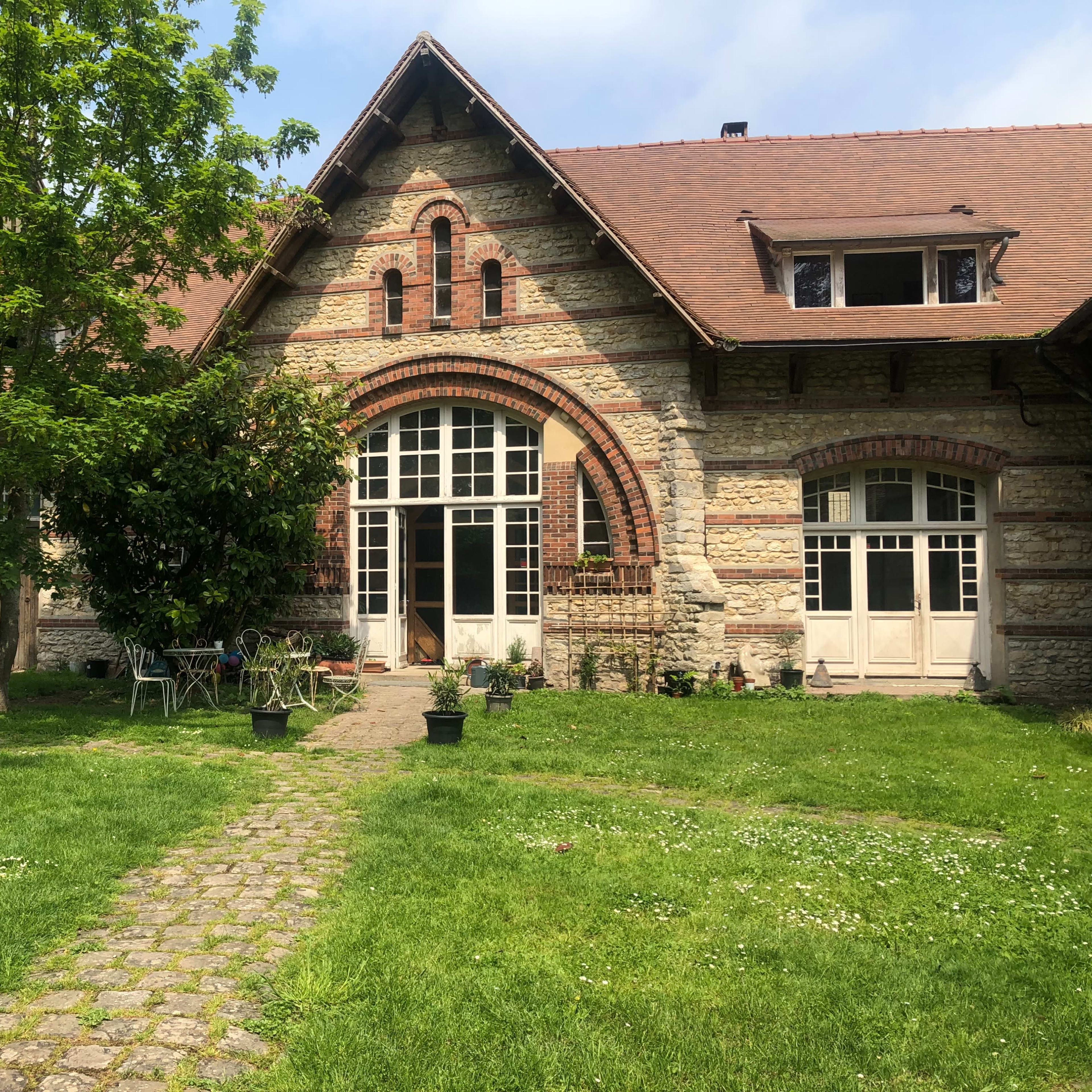 A stone house with arched windows and a sloped roof is surrounded by greenery and a stone pathway leading to the entrance.