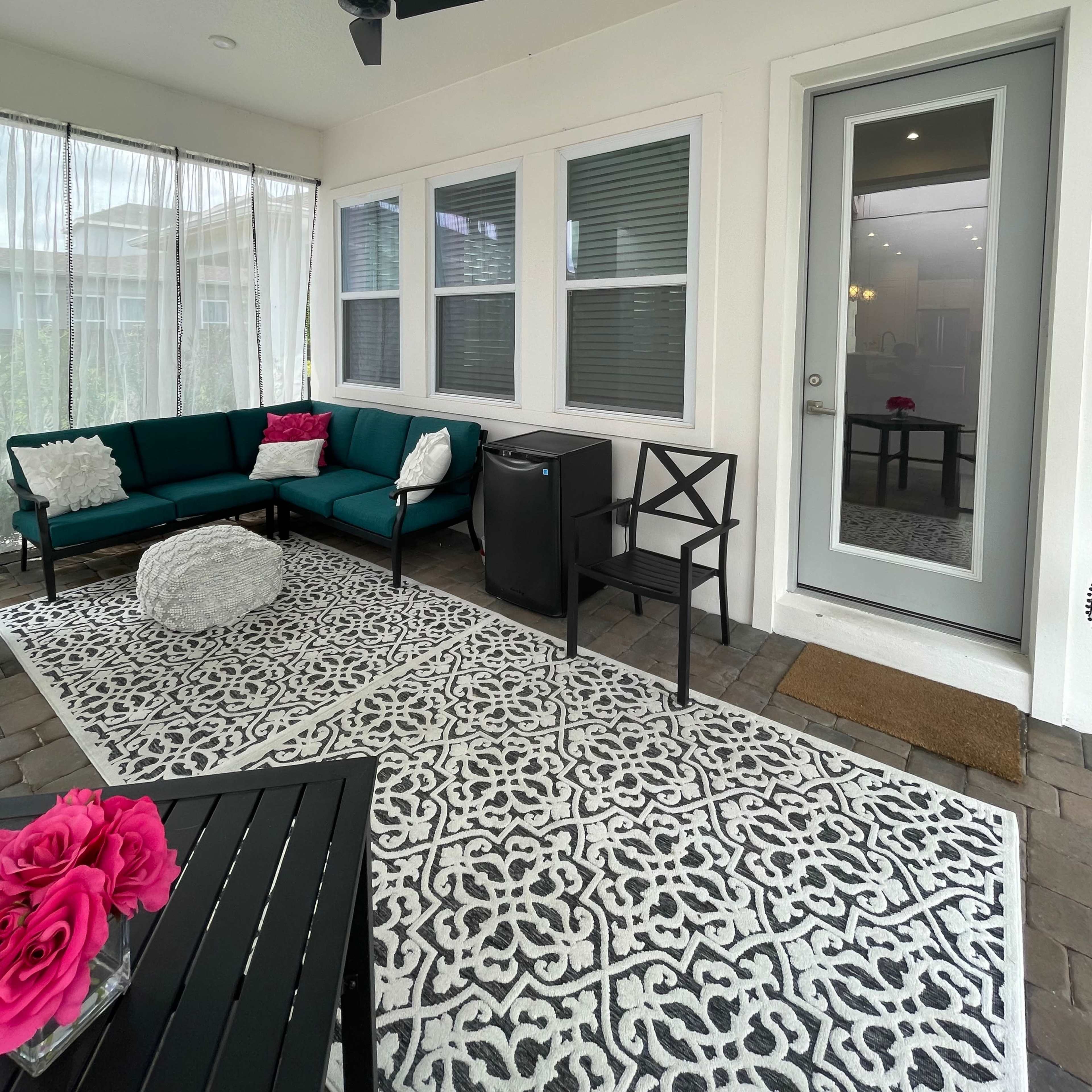 A covered patio area features a green sofa with pink cushions, a round pouf, a small table, and a door leading into an interior space.