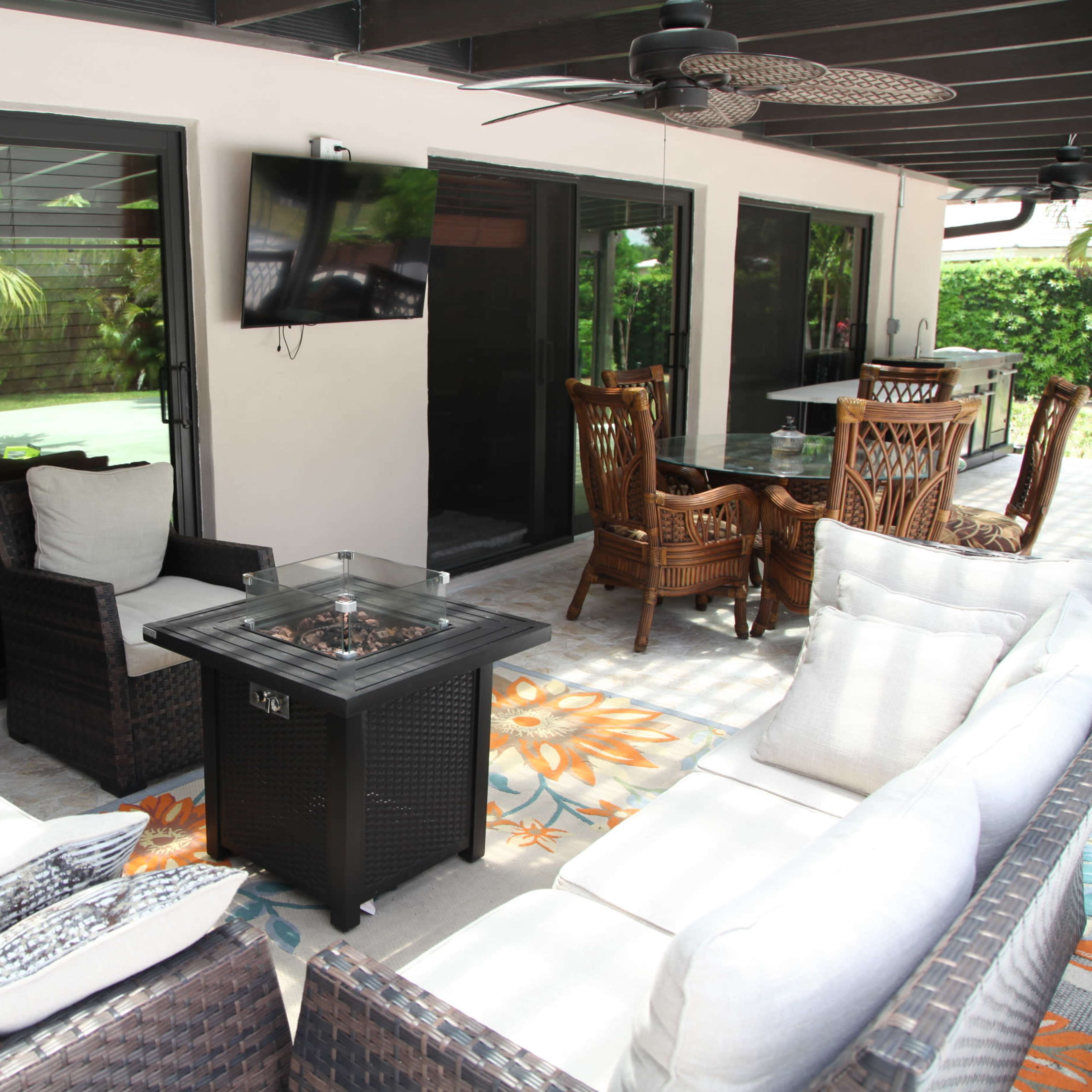 A covered patio area with wicker furniture arranged around a fire pit, a television mounted on the wall, and a dining area in the background.