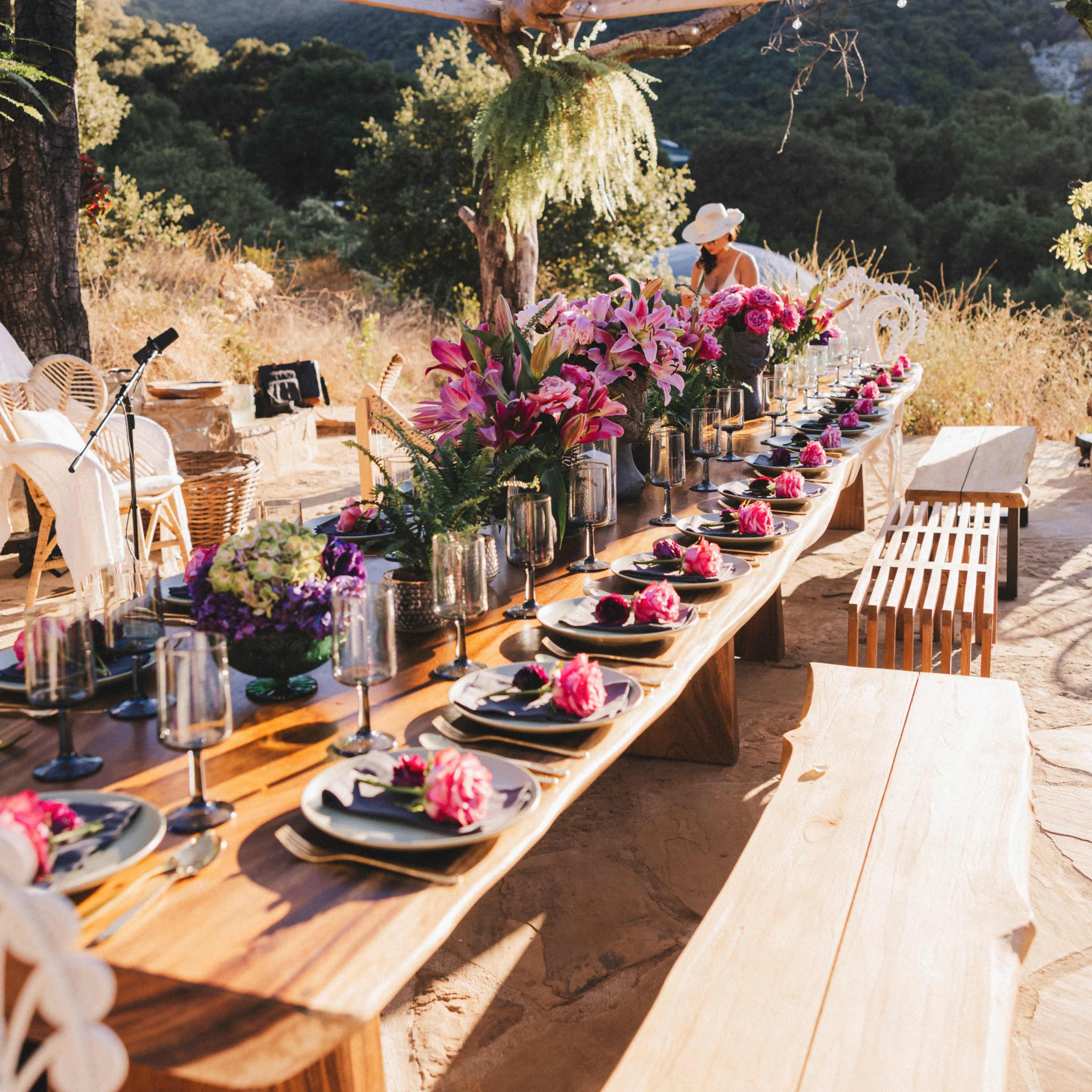 A long wooden table is set outdoors with elegant place settings and vibrant floral arrangements under a canopy of bare branches.
