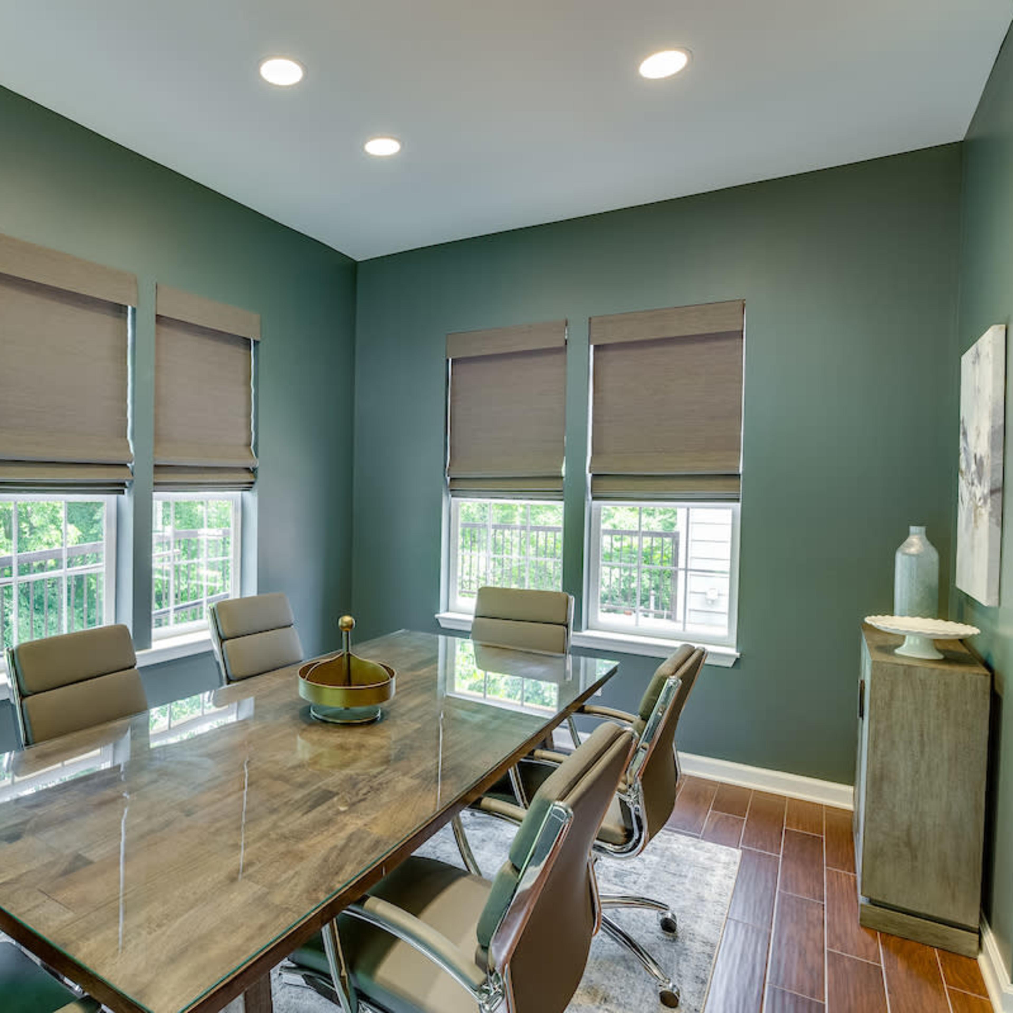 A modern conference room with a glass table surrounded by silver chairs, featuring large windows covered with brown shades.