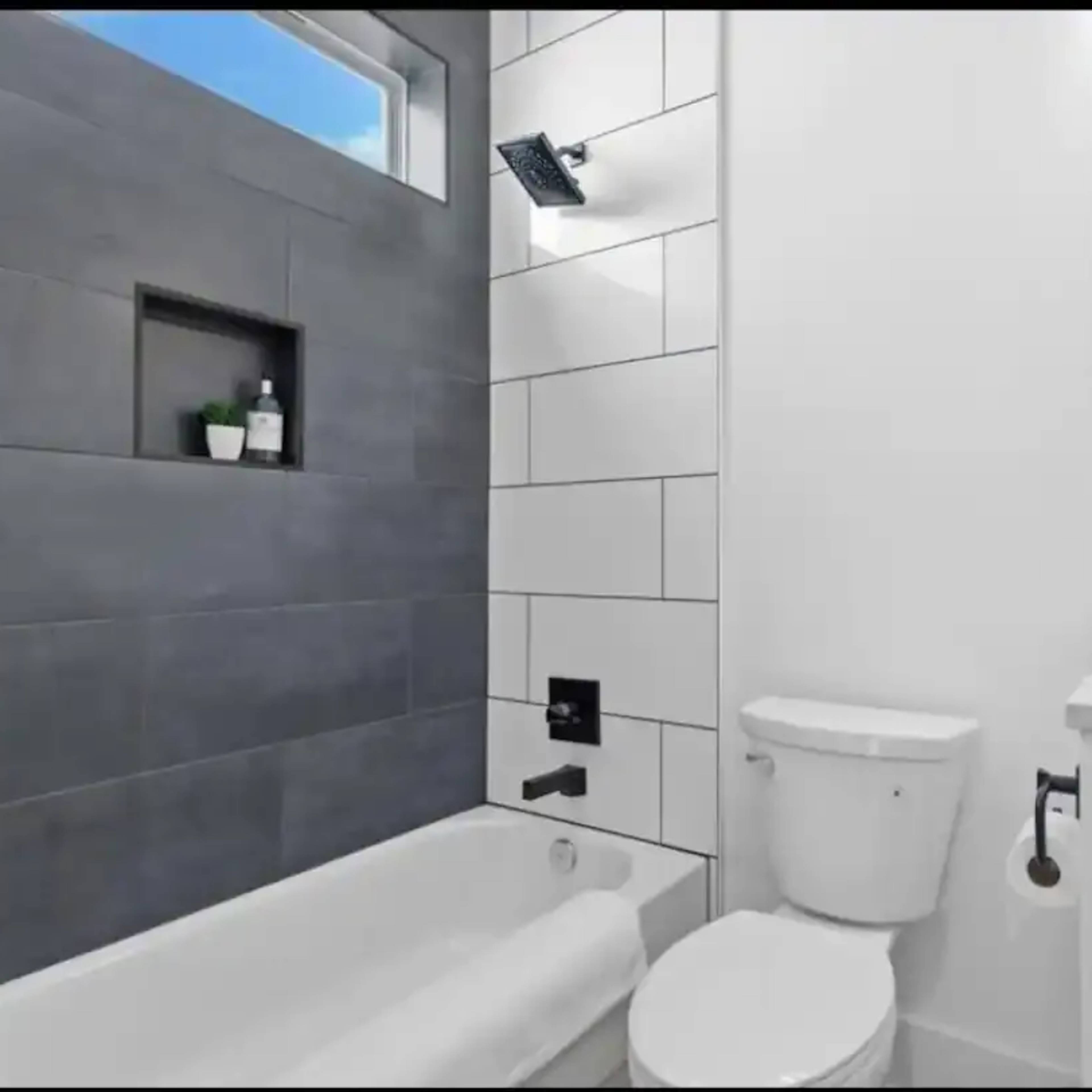 A modern bathroom featuring a white bathtub, a toilet, a wall with gray and white tiles, and a small shelf holding plants.