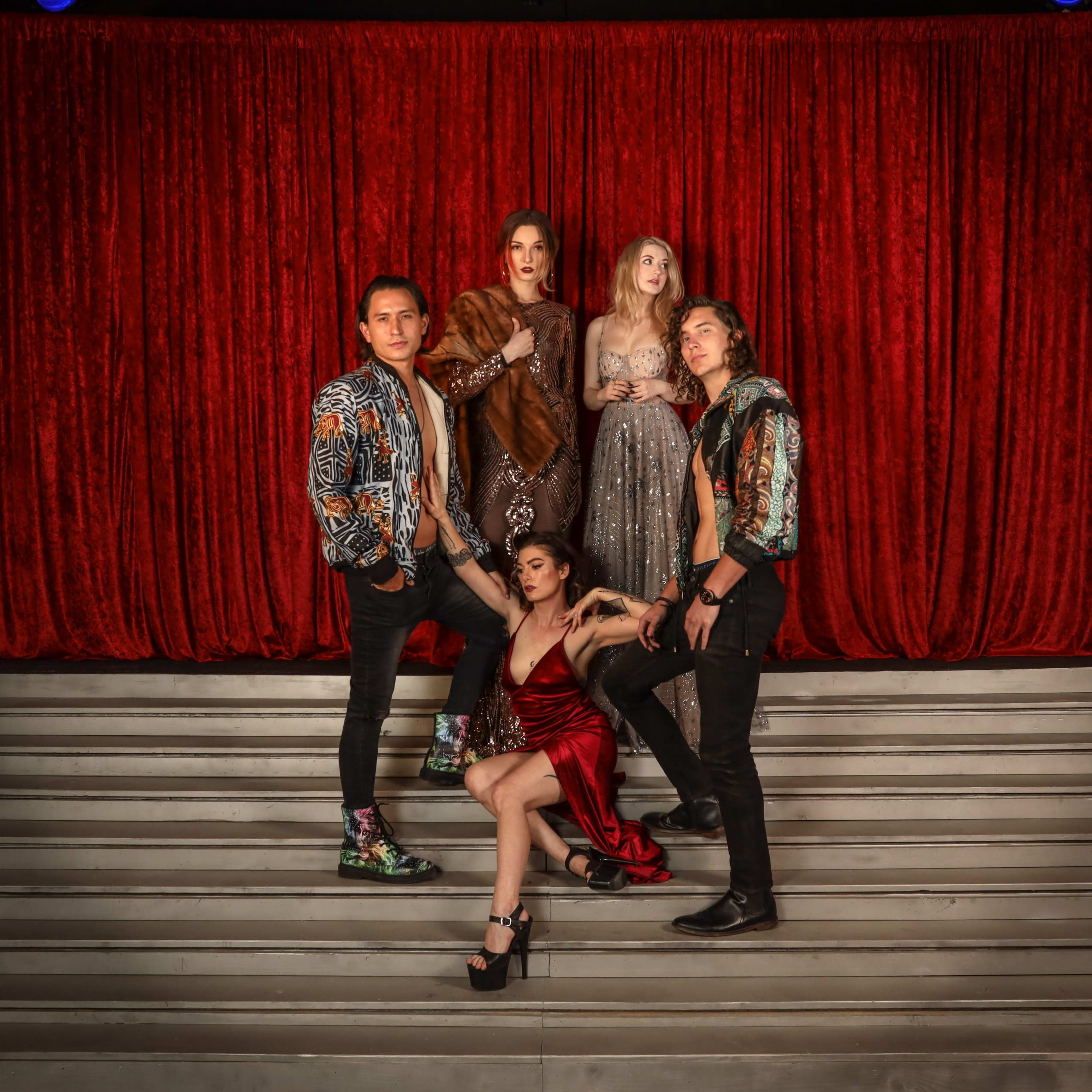A group of five individuals poses on a staircase in front of a red curtain, showcasing a mix of stylish outfits.