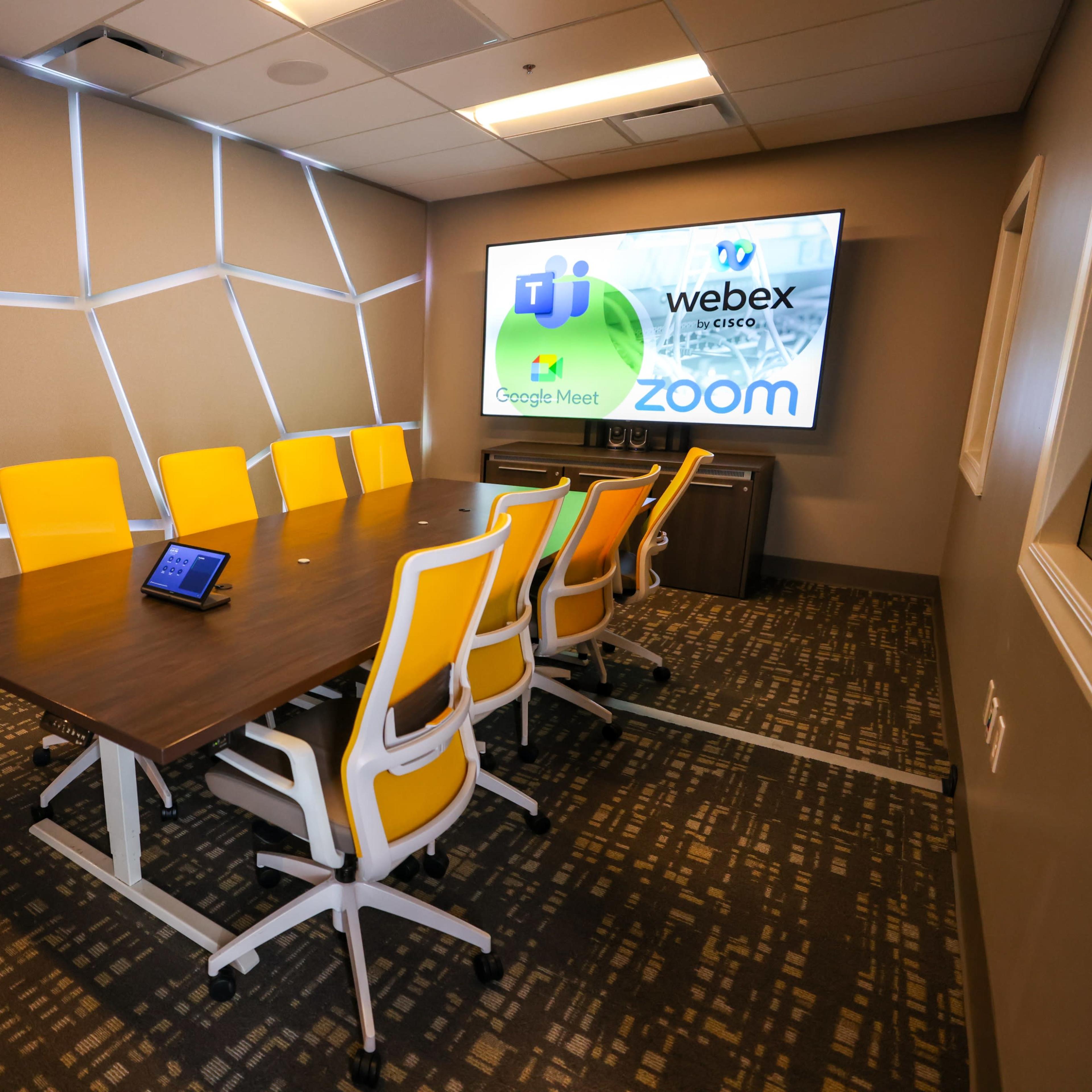 A modern conference room features a long wooden table with yellow chairs and a screen displaying logos for various video conferencing platforms.