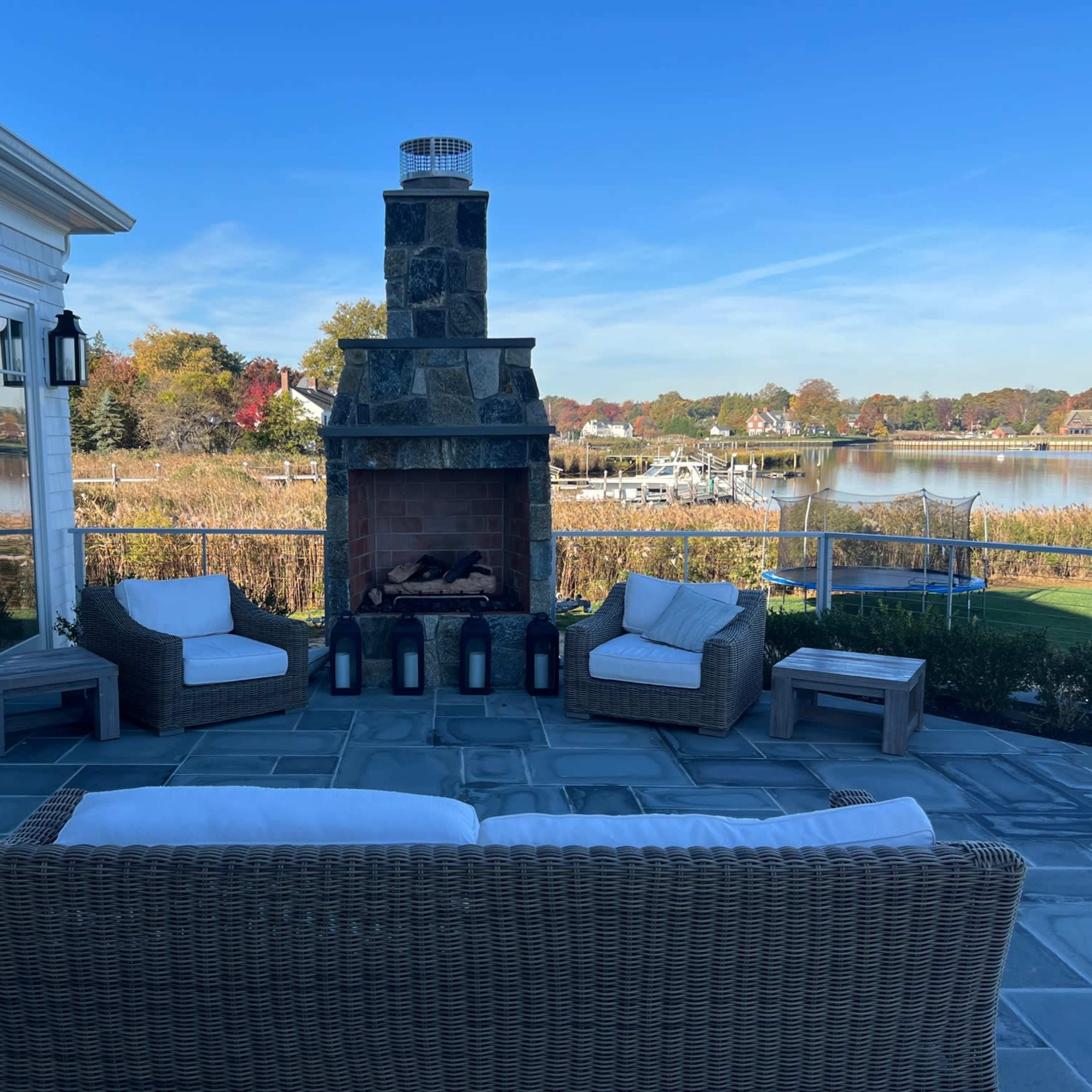 An outdoor seating area with a stone fireplace, wicker furniture, and a view of a waterfront landscape.