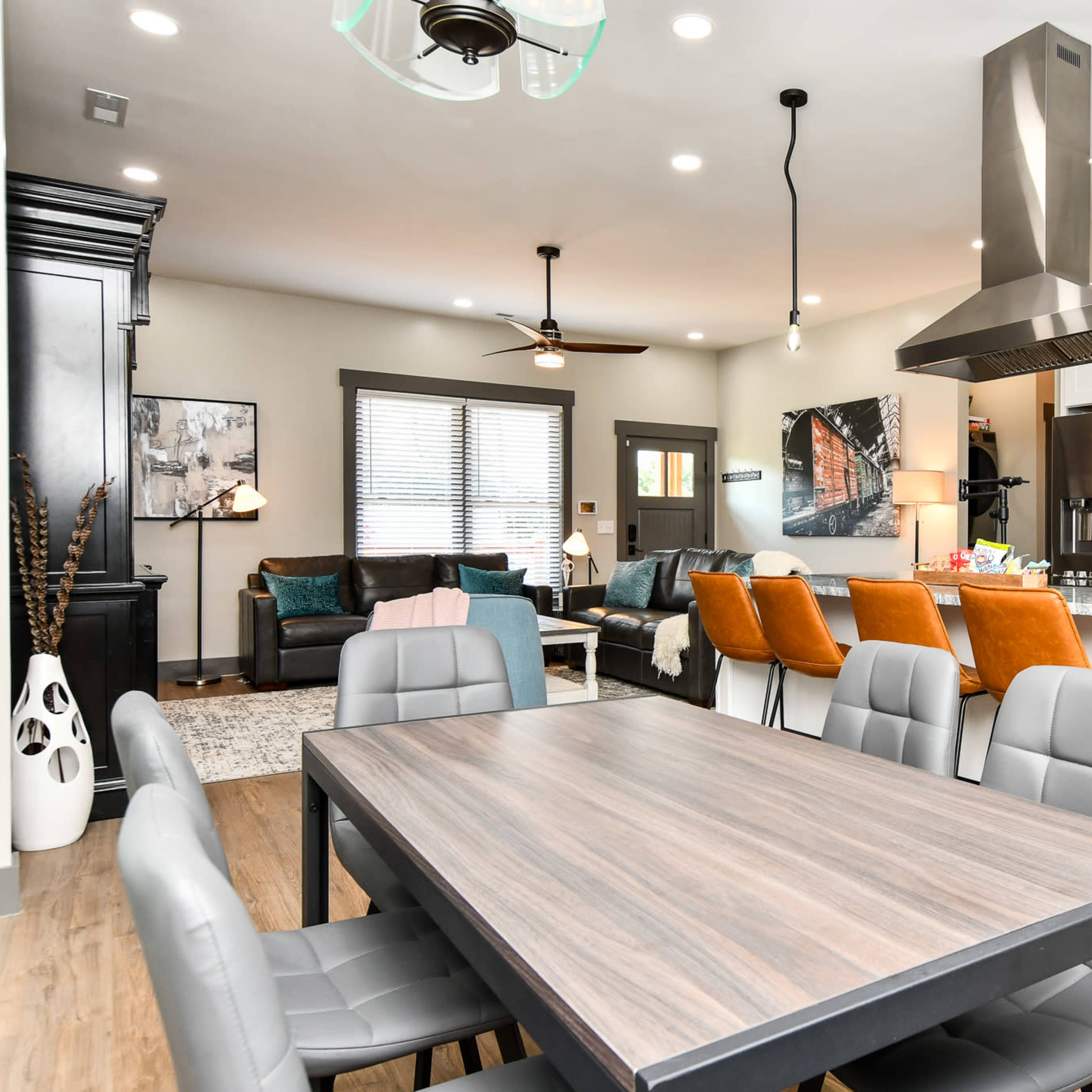 A modern open-concept living and dining area with a dining table in the foreground, a kitchen with stainless steel appliances, and a cozy seating arrangement in the background.