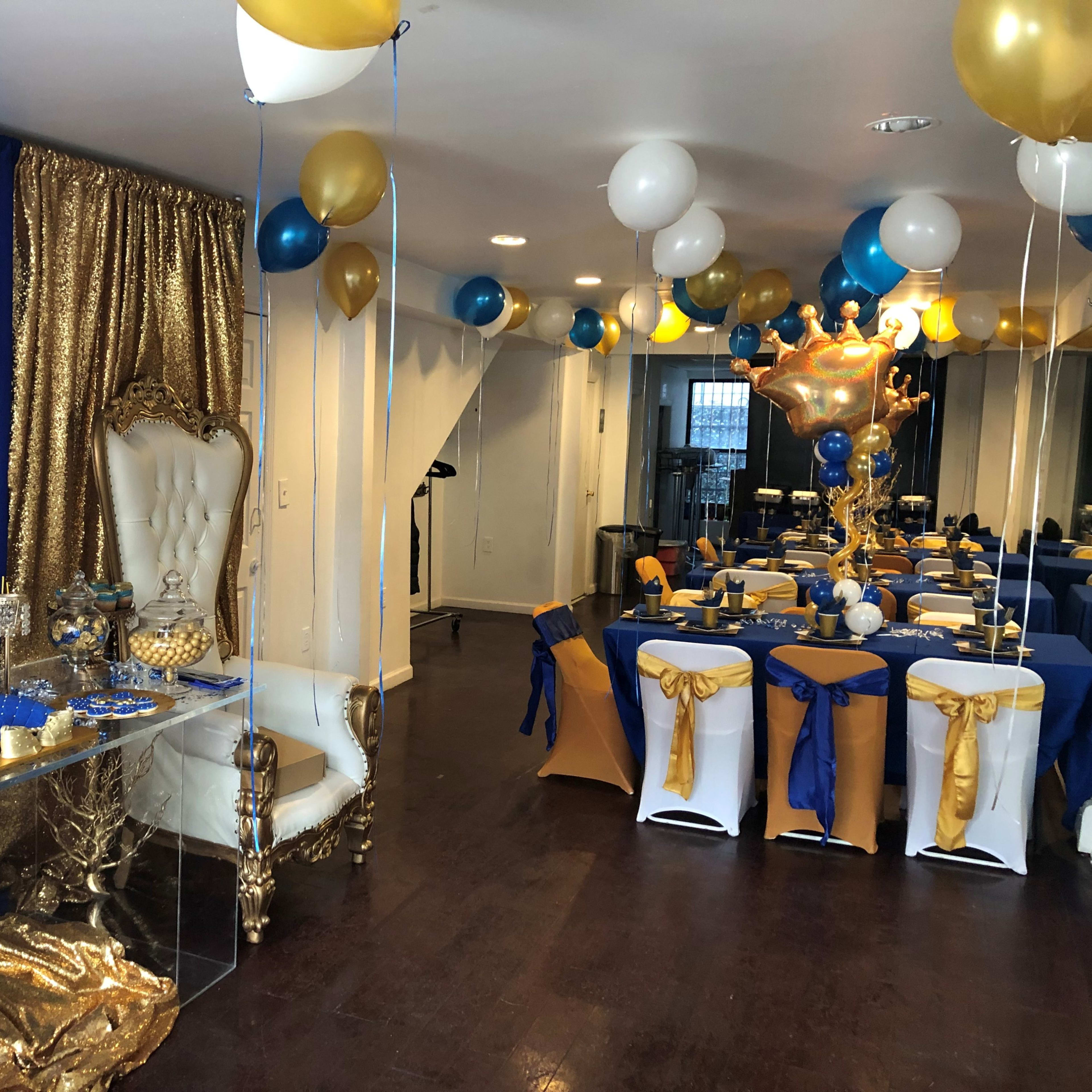 A decorated event space featuring a dessert table with golden drapery, white and blue balloons, and elegantly set tables with blue and gold tablecloths.