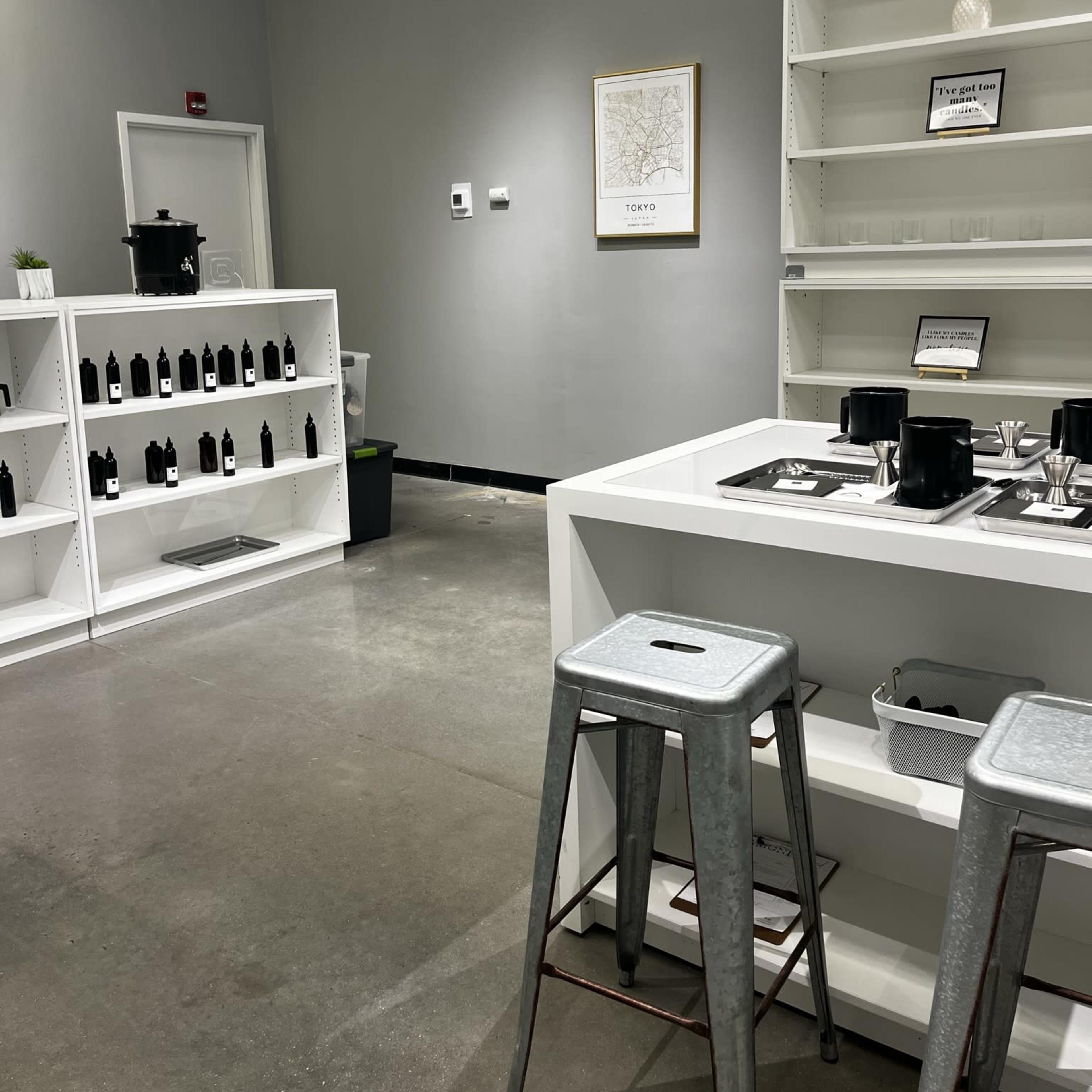 A minimalist indoor space with white shelves containing black bottles, a light-colored wall featuring a framed map, and a central white table with metal stools.