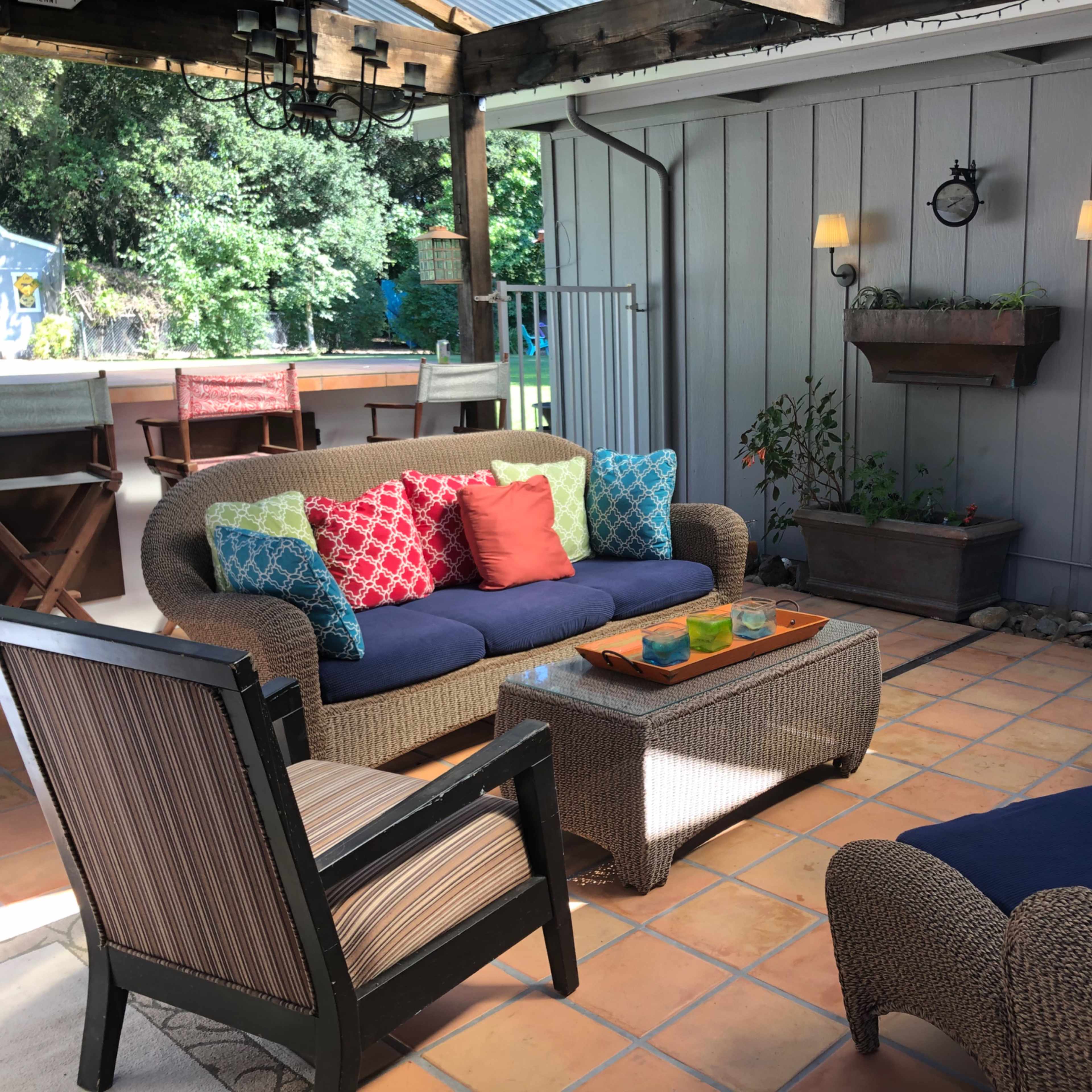 The scene features a patio area with a woven sofa adorned with colorful cushions, a wooden coffee table with decorative items, and surrounding chairs on a tiled floor.