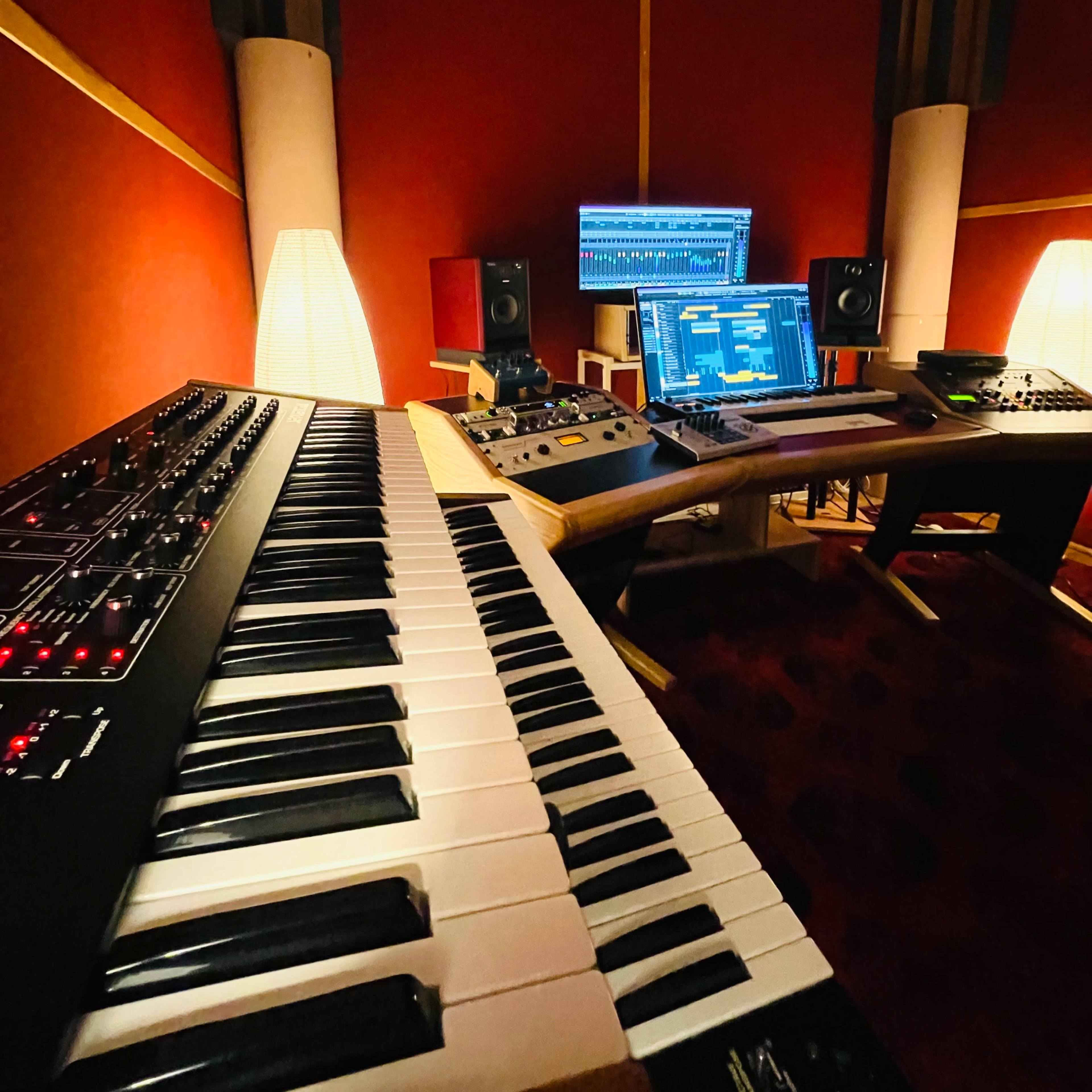 A music production studio with a keyboard, computer monitor, and audio equipment against a red wall.