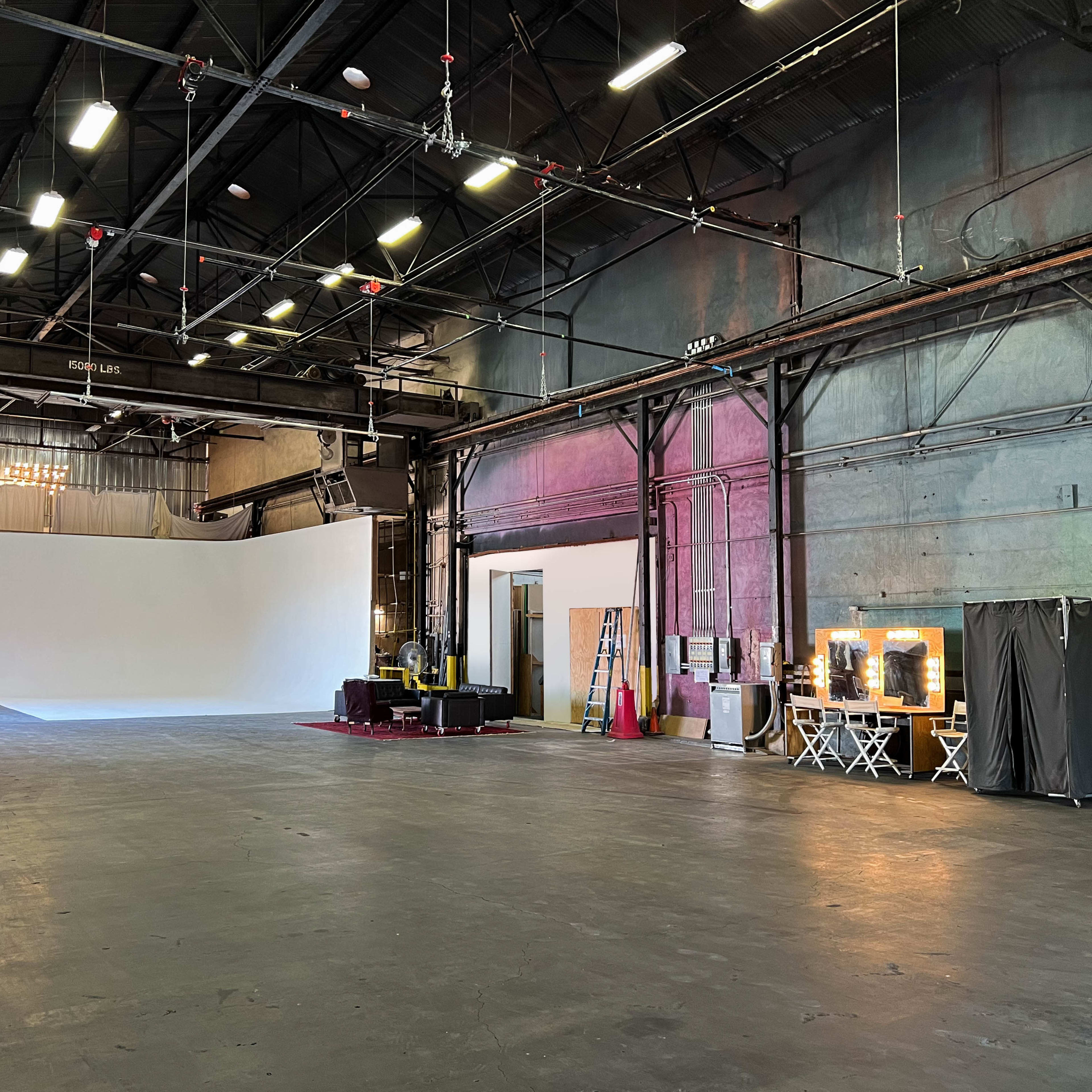A spacious industrial interior with a large open area, white backdrop, and various sets of lighting and makeup stations along one side.