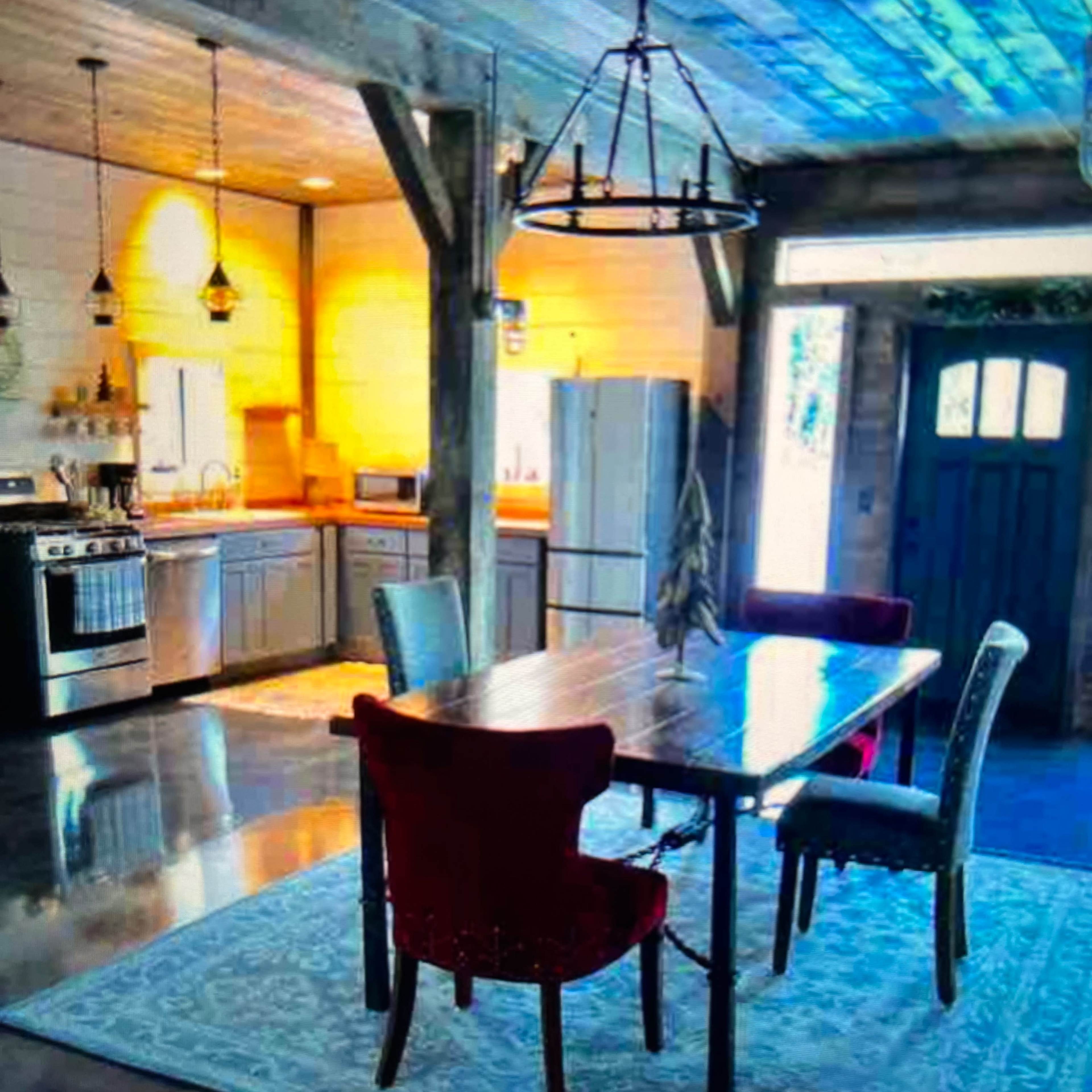A modern kitchen features a wooden dining table surrounded by chairs, with stainless steel appliances and large windows allowing natural light to enter.