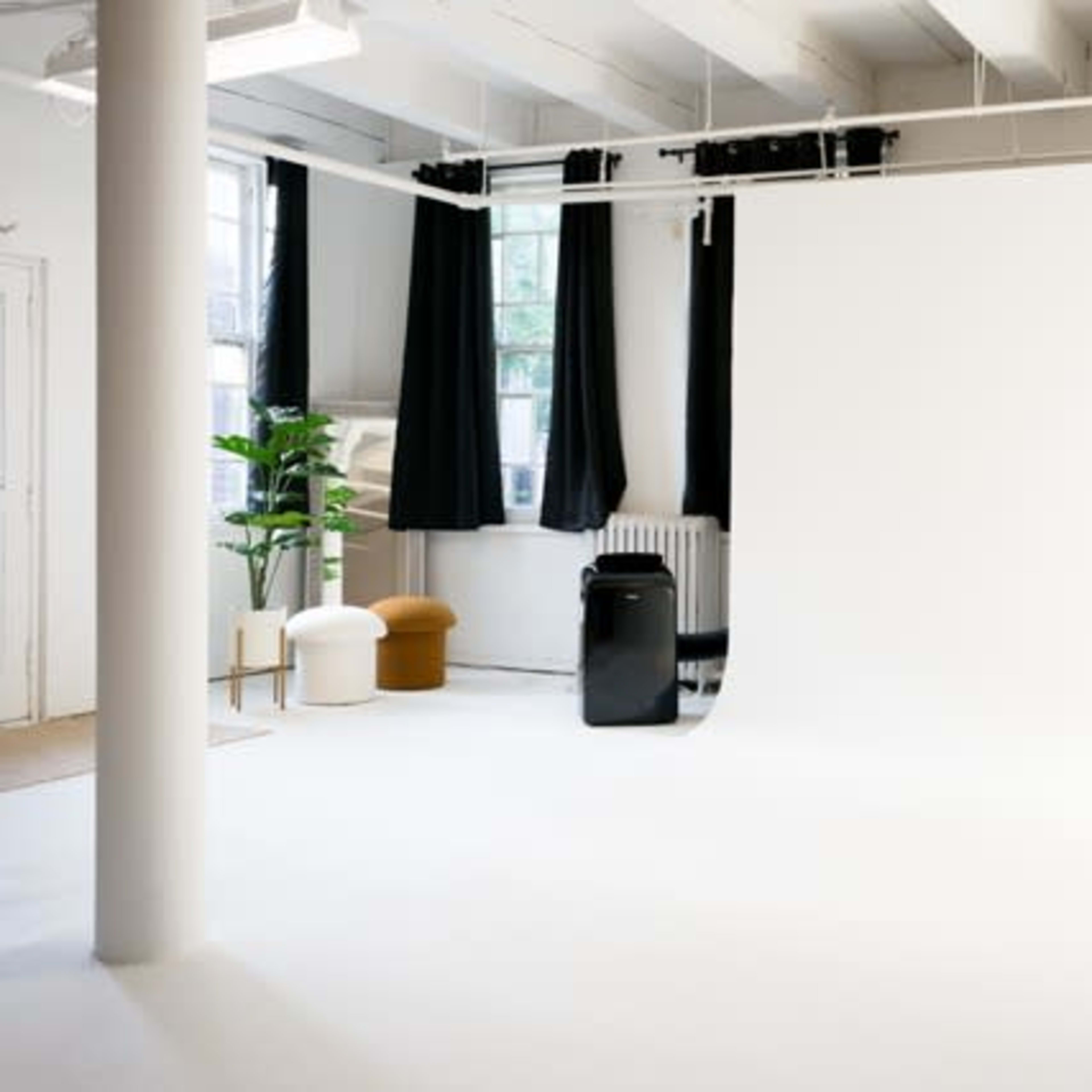 A spacious studio with a white backdrop, a green plant, and a few pieces of furniture under a bright ceiling.