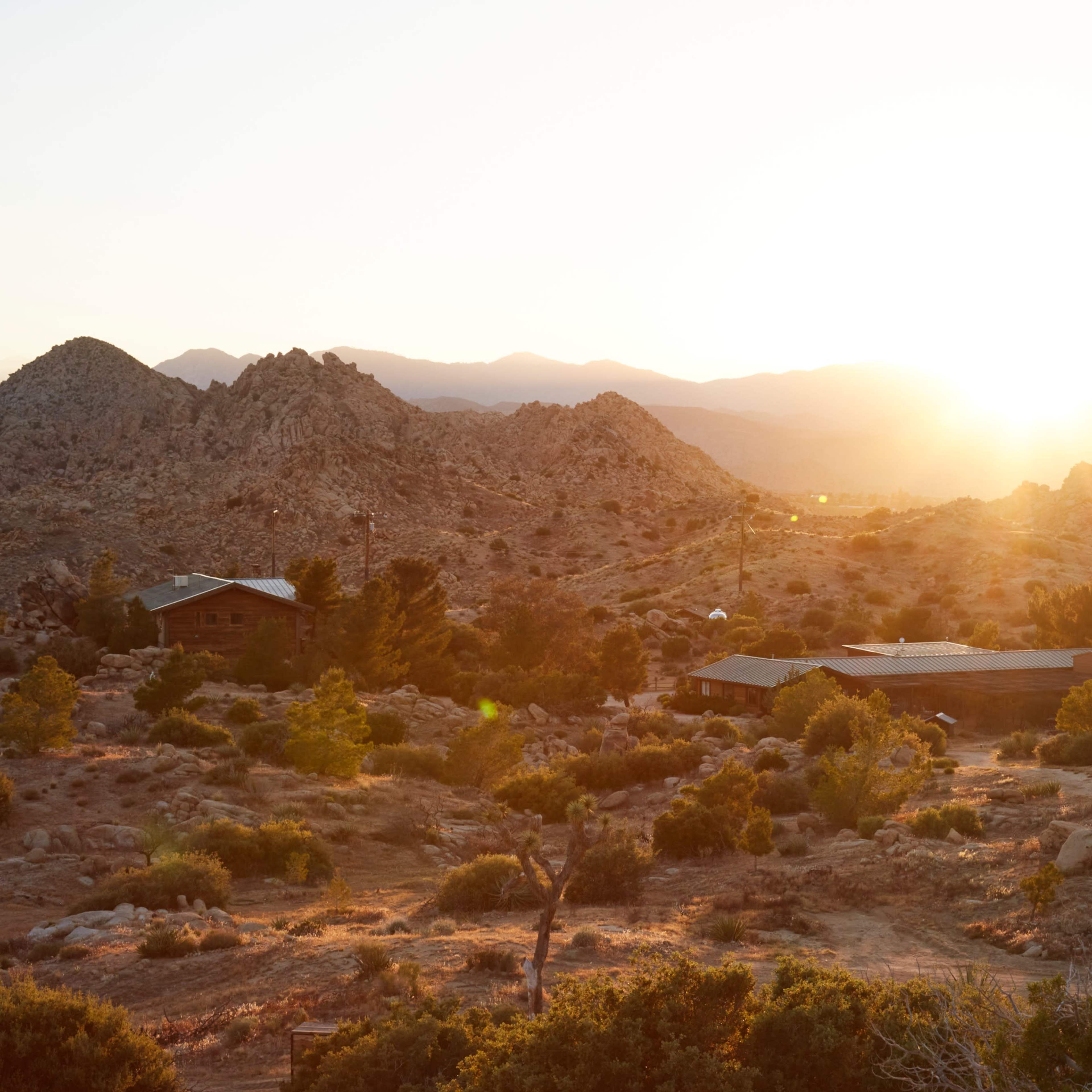 The sun rises over a rocky landscape dotted with sparse vegetation and a cluster of wooden buildings.