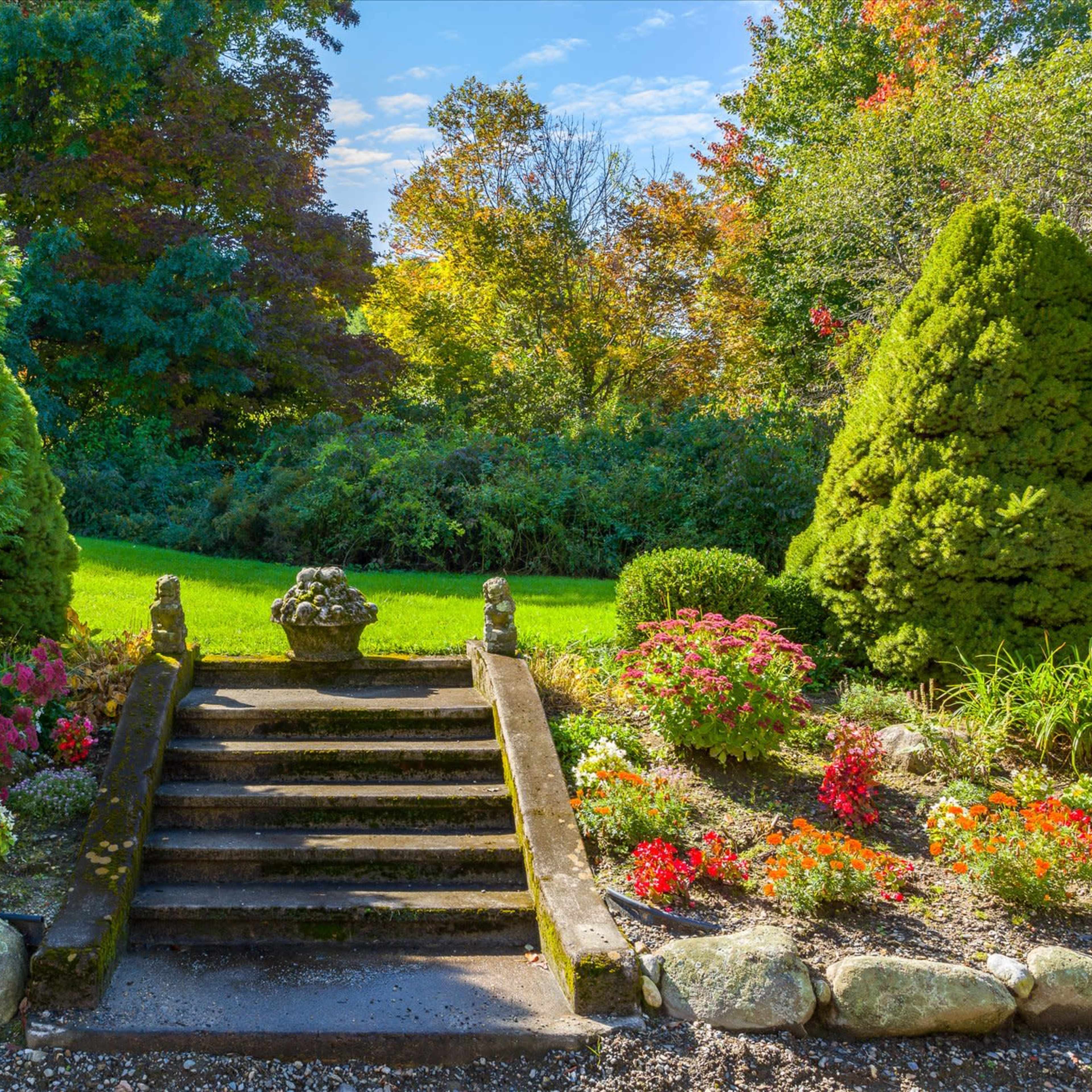 A landscaped garden features stone steps leading to a flower bed, flanked by neatly trimmed shrubbery and colorful flower arrangements.