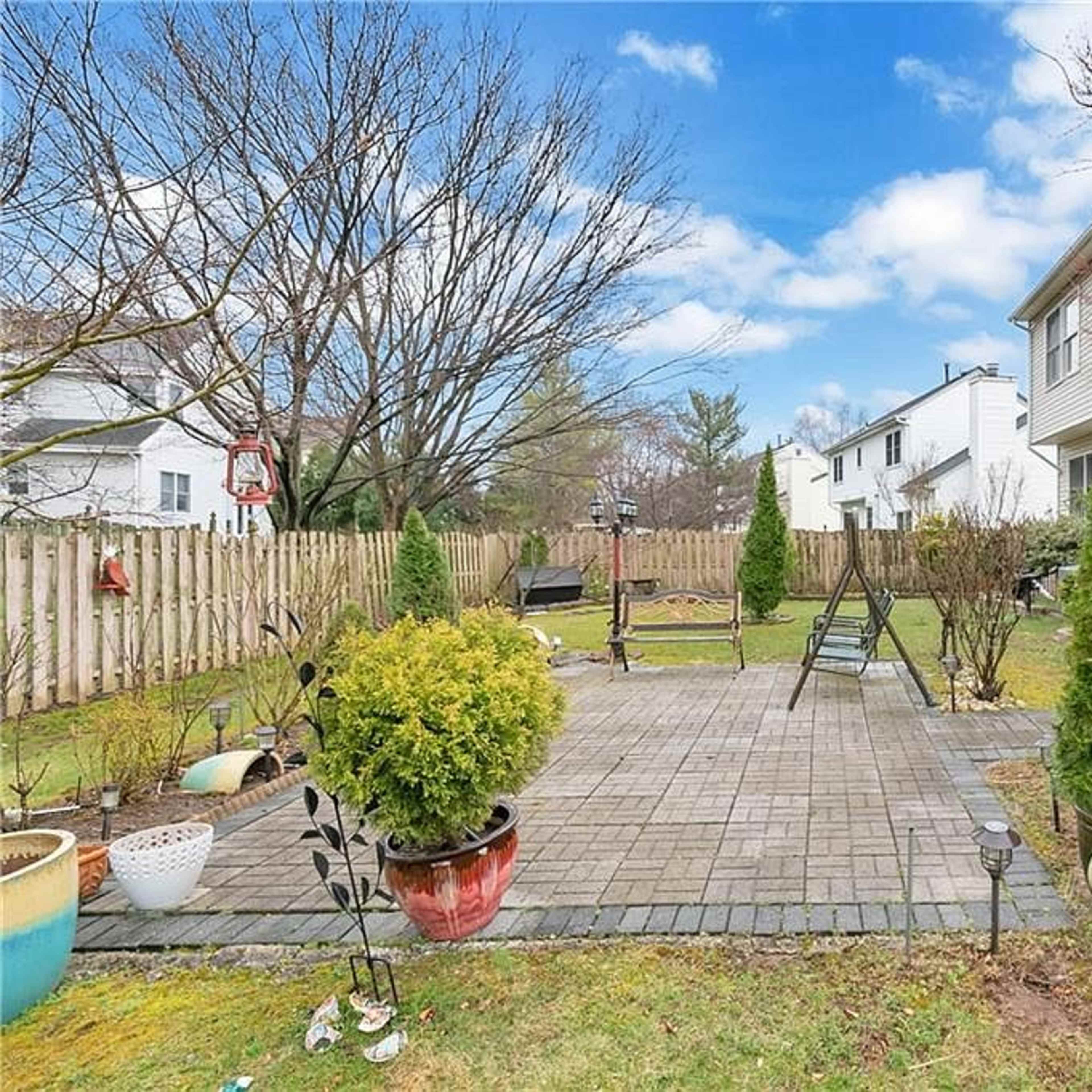 A backyard with a stone patio, several potted plants, and a wooden deck leading to a house, surrounded by a wooden fence and grass.