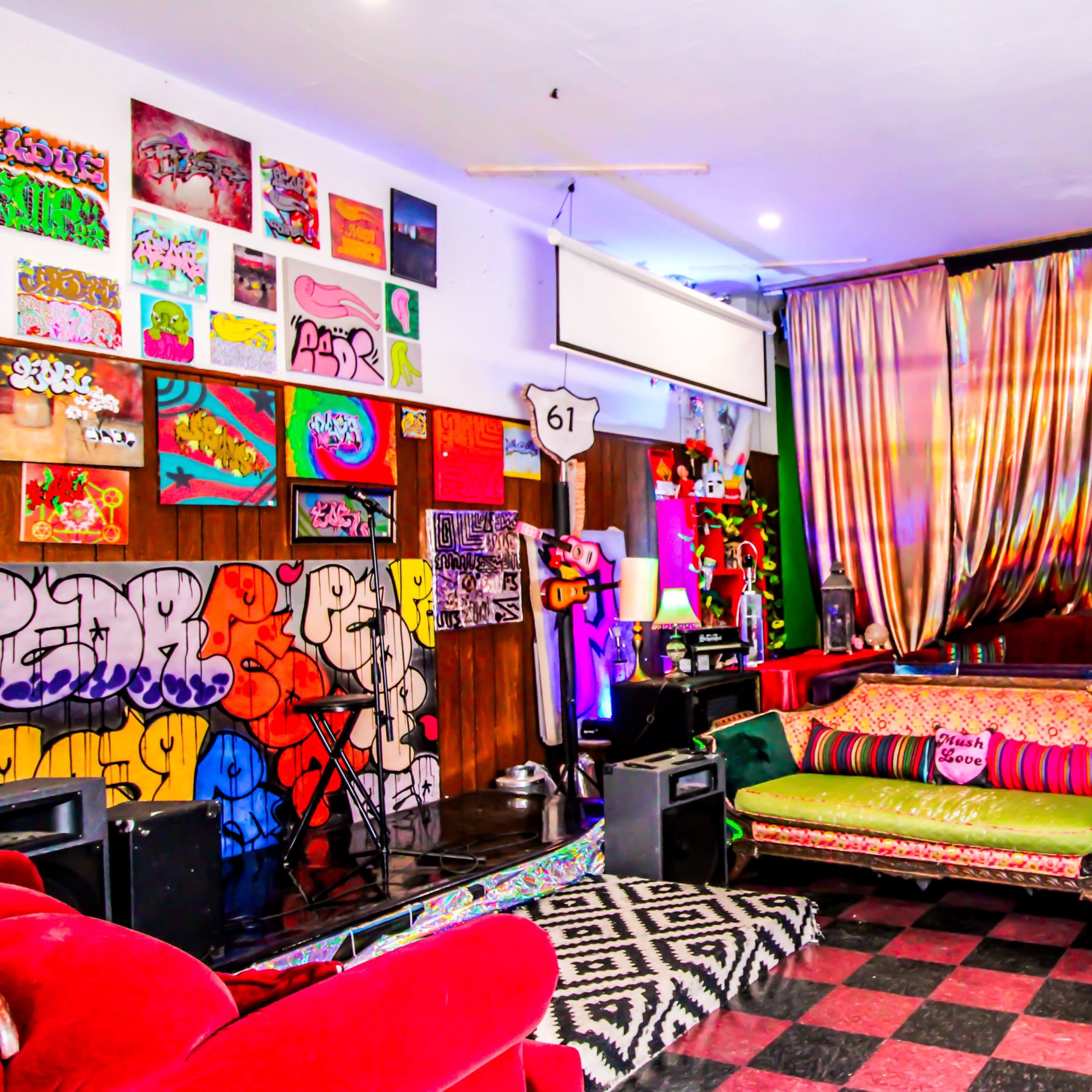 A vibrant, eclectic lounge area featuring colorful art on the walls, various seating options, and a stage-like setup at one end.