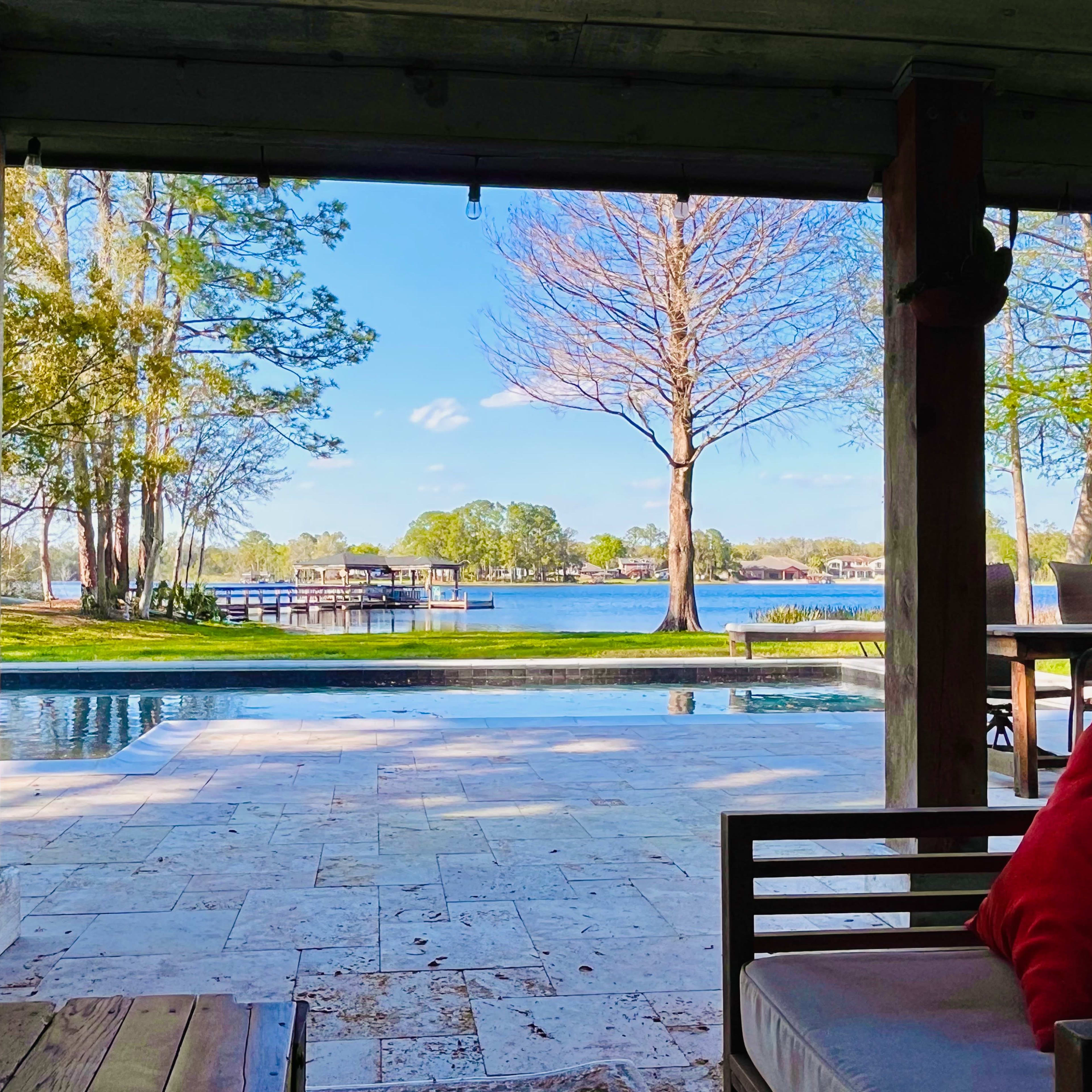 A view from a covered porch overlooking a pool and lake surrounded by trees.