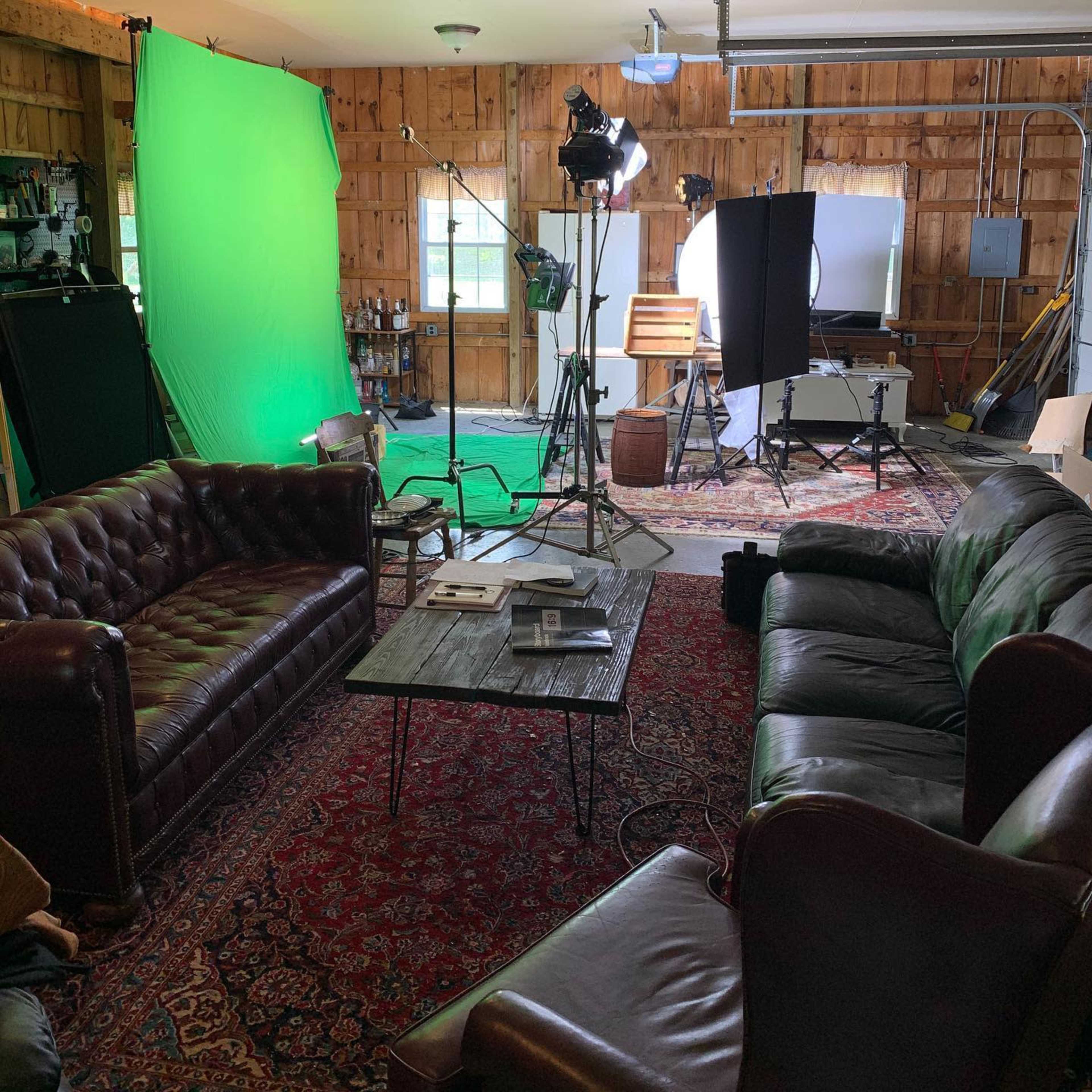 A studio setup with green screens, lighting equipment, and two brown sofas arranged around a central coffee table on a patterned rug.