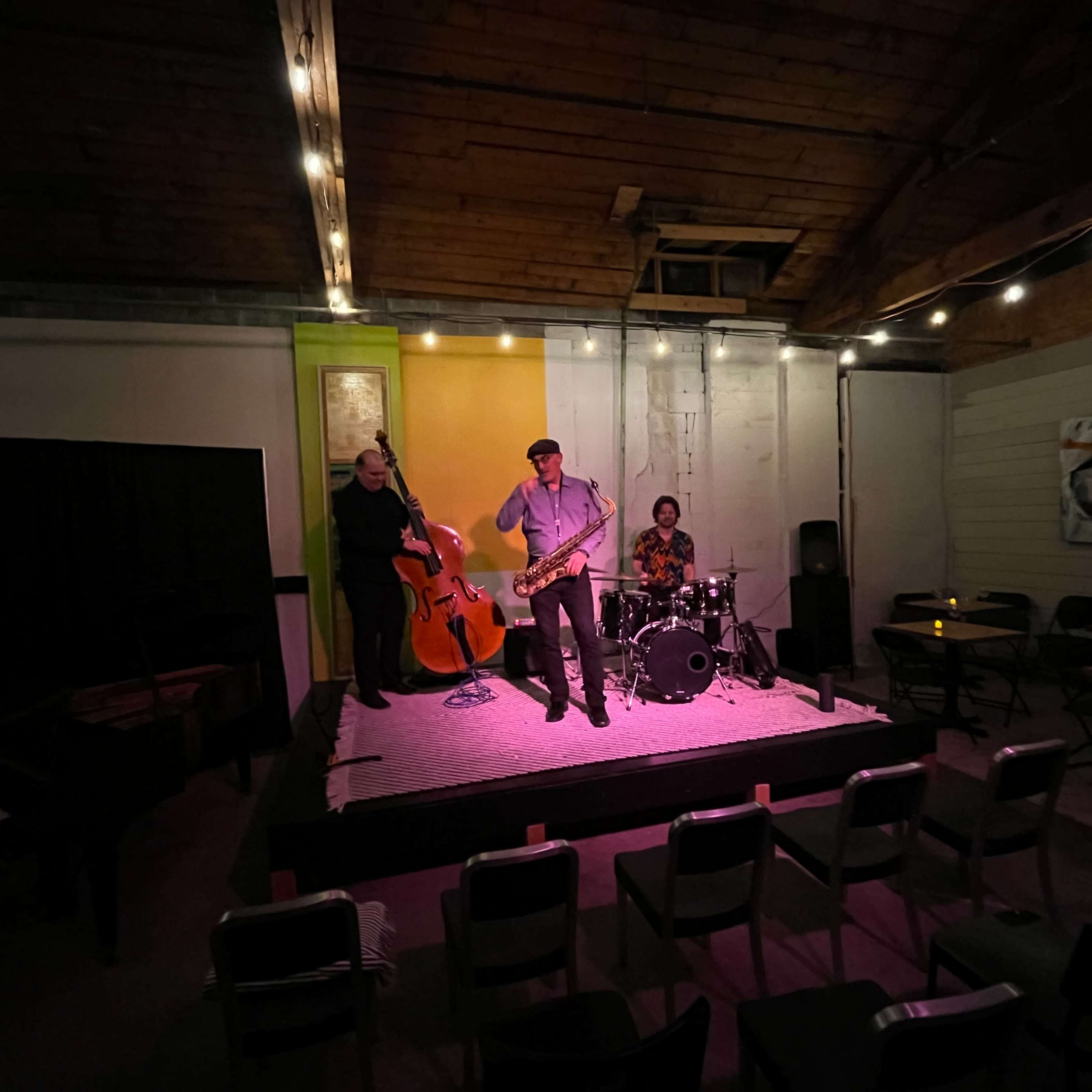 A jazz trio performs on a small stage in a dimly lit room, featuring a saxophonist, a double bassist, and a drummer.