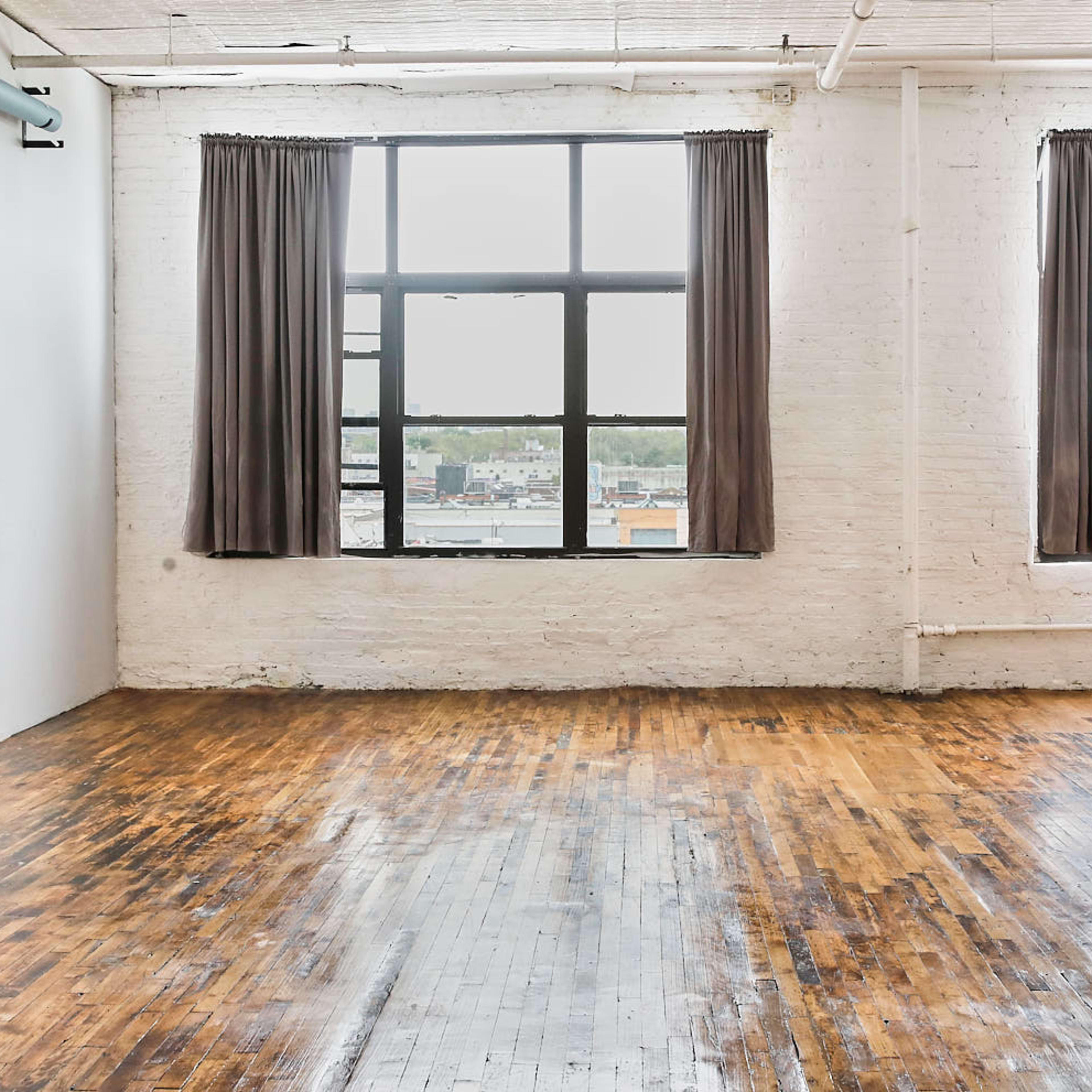 A spacious, empty room with large windows, wooden floors, and white brick walls.