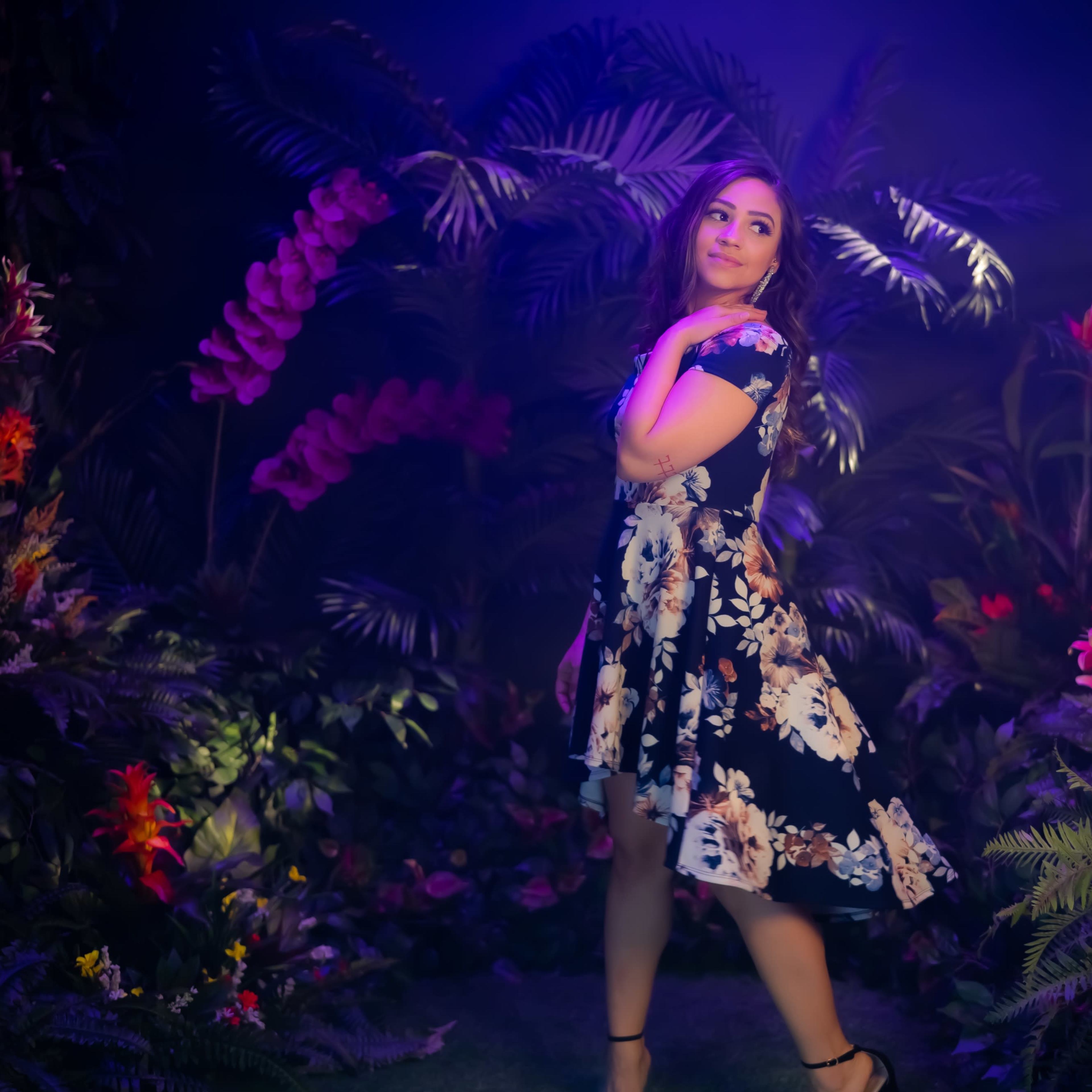 A woman poses elegantly in a floral dress within a vibrant, plant-filled environment illuminated by colorful lights.