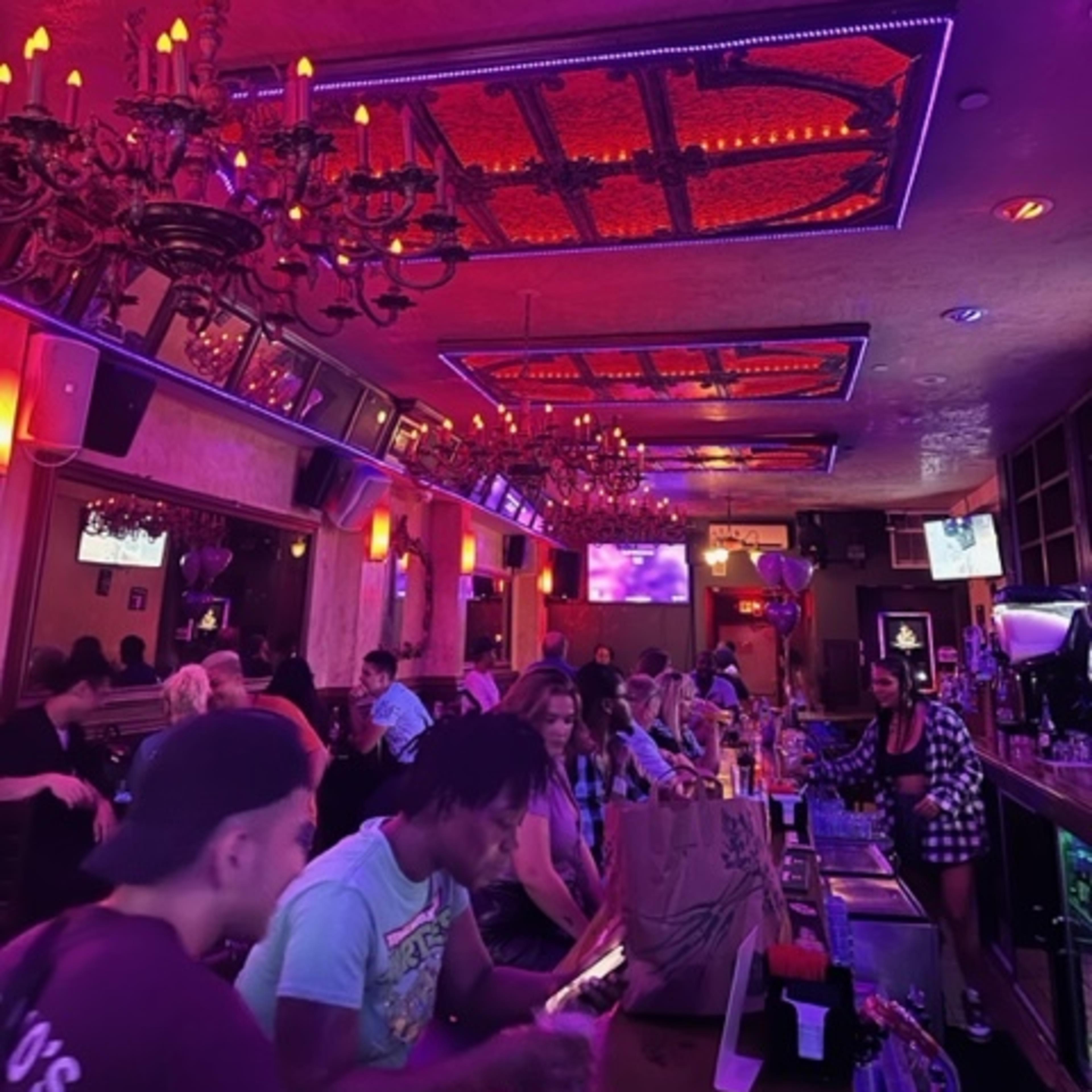 A lively bar scene features patrons sitting at the counter and tables, illuminated by purple and red lights, with ornate chandeliers hanging from the ceiling.