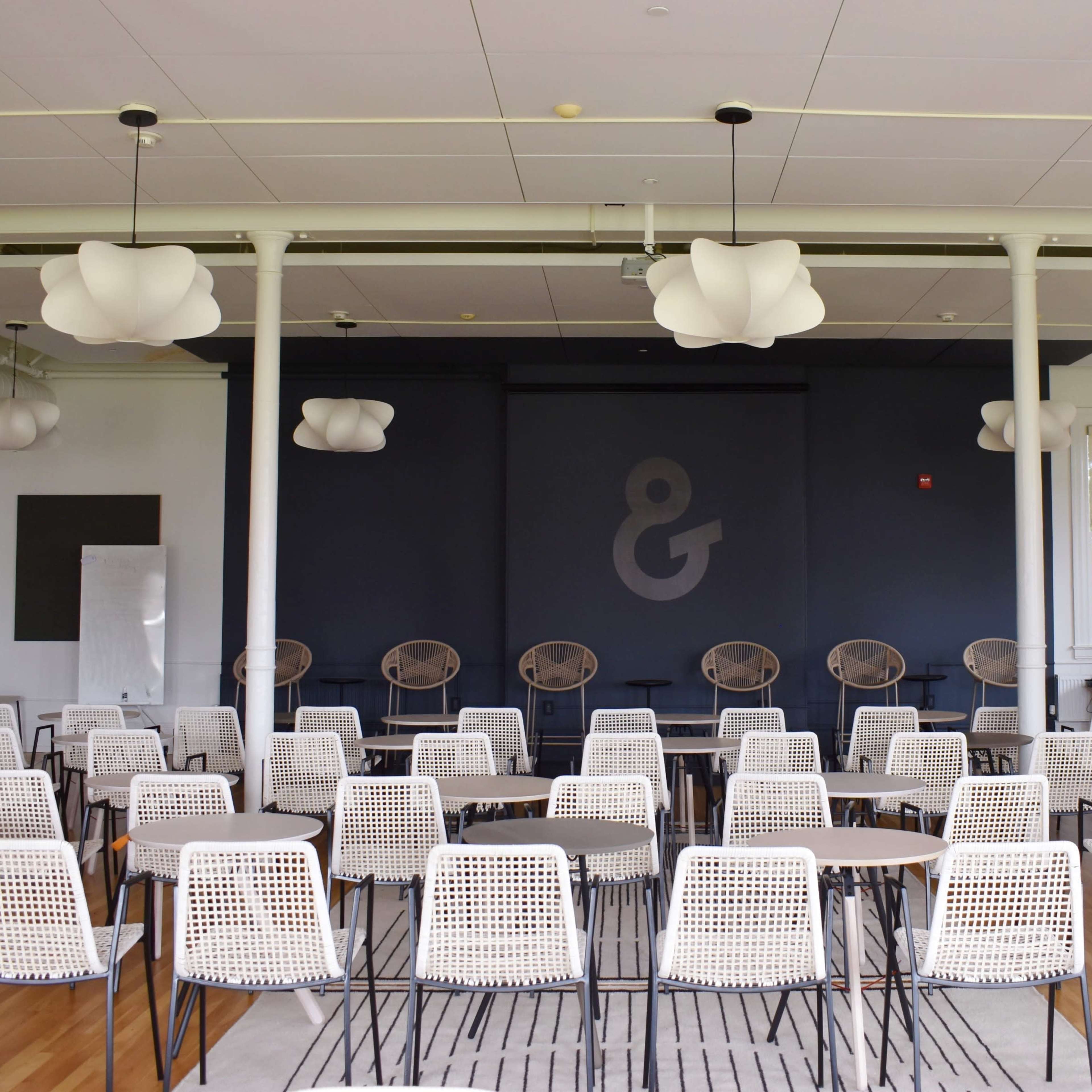 A modern meeting room set up with rows of chairs and tables arranged facing a large dark wall, adorned with a stylized ampersand symbol, and featuring distinctive pendant lights above.
