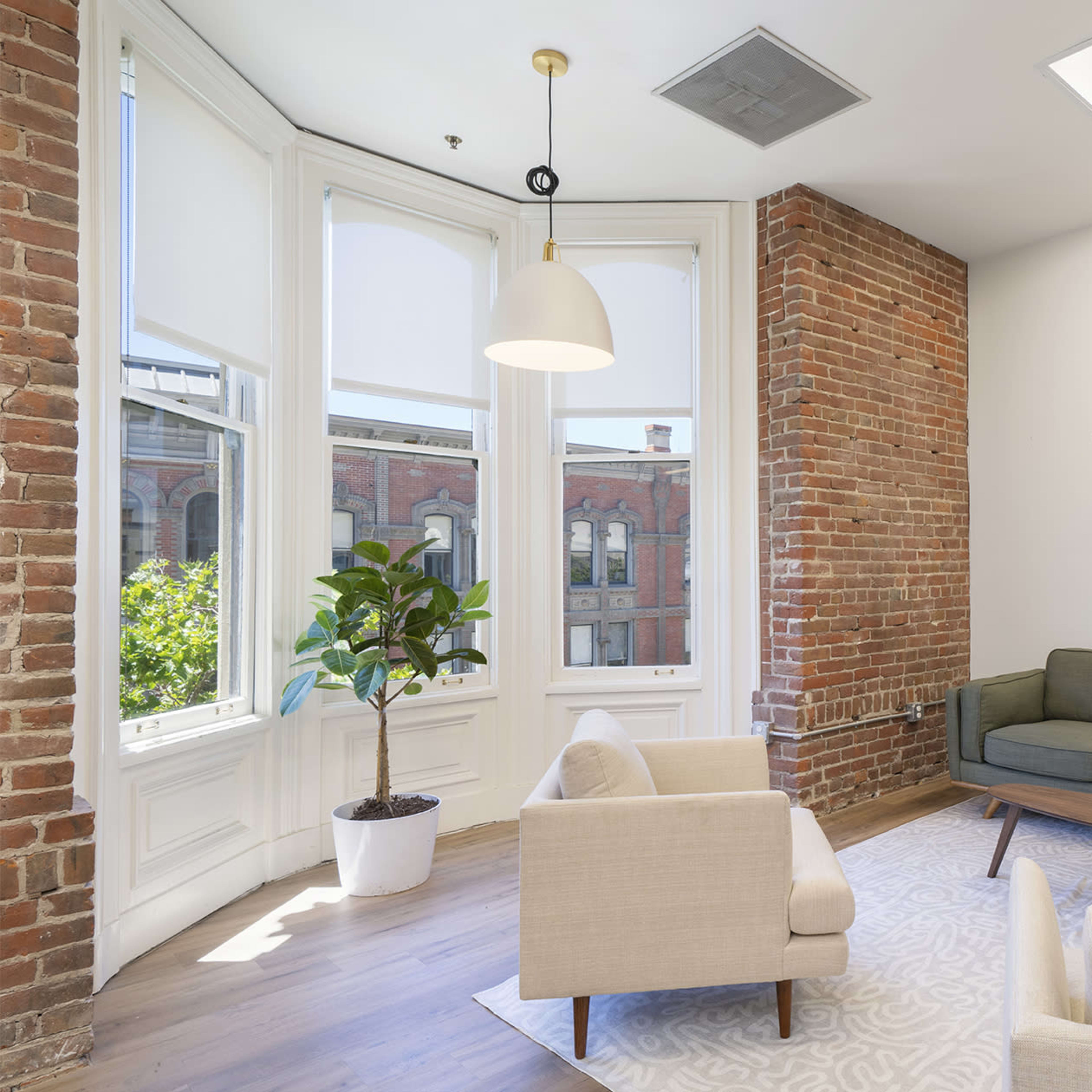 A cozy sitting area featuring a green sofa, two light-colored armchairs, a wooden coffee table, and a potted plant, with large windows letting in natural light and exposed brick walls.