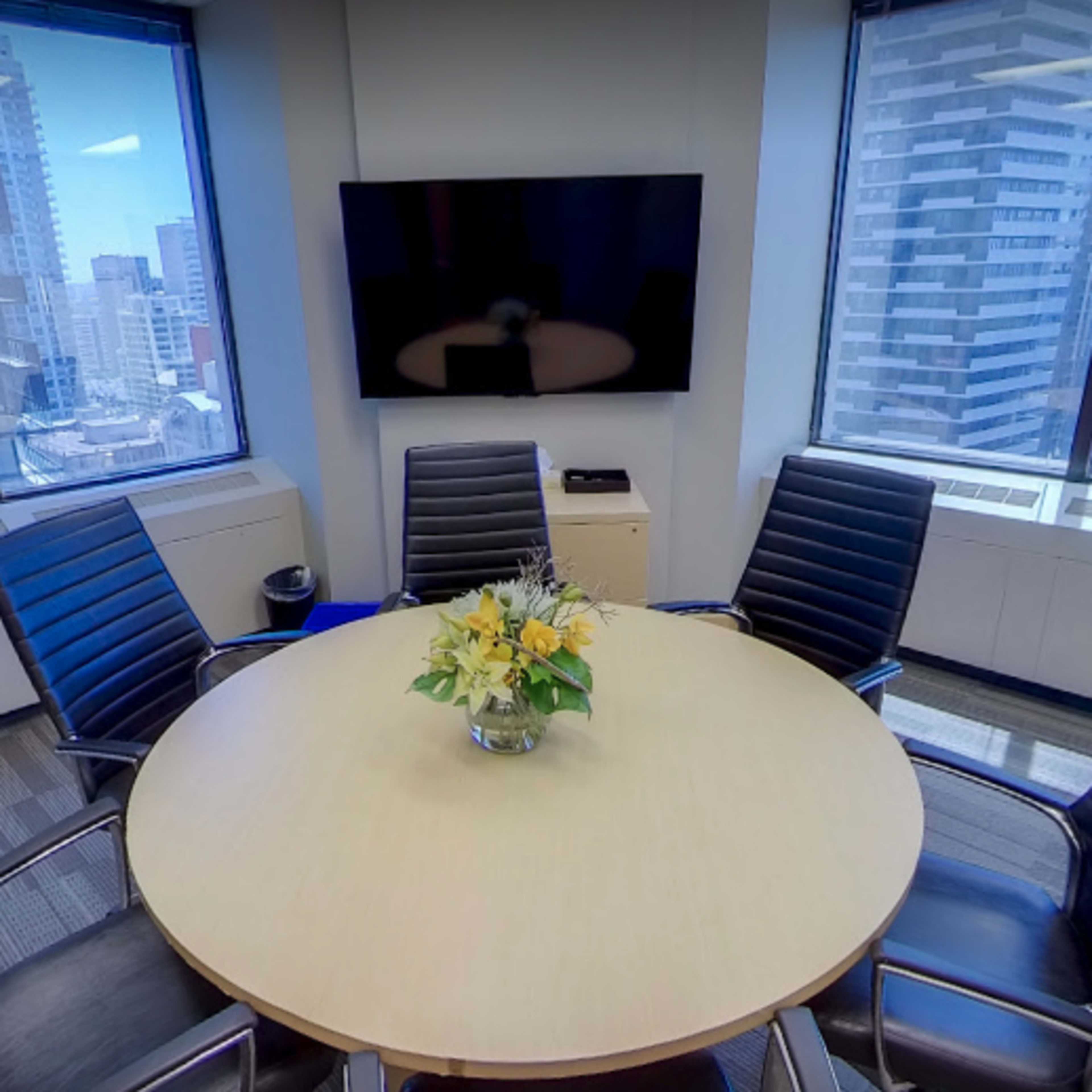 A conference room features a circular table surrounded by black chairs, large windows providing city views, and a wall-mounted television.